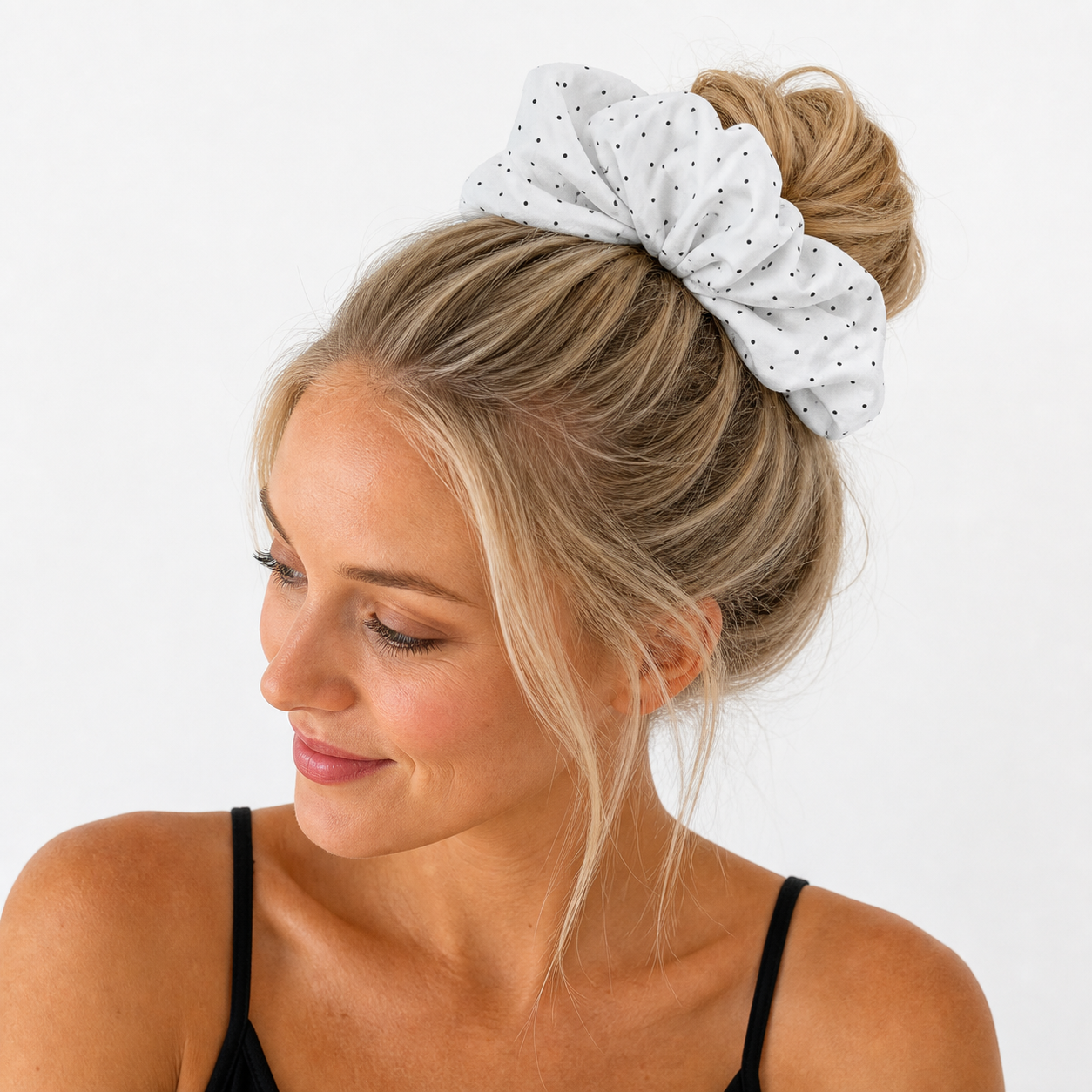 Blonde woman wearing a white black polka dot scrunchie in a high messy bun, side profile, black camisole top, studio white background.