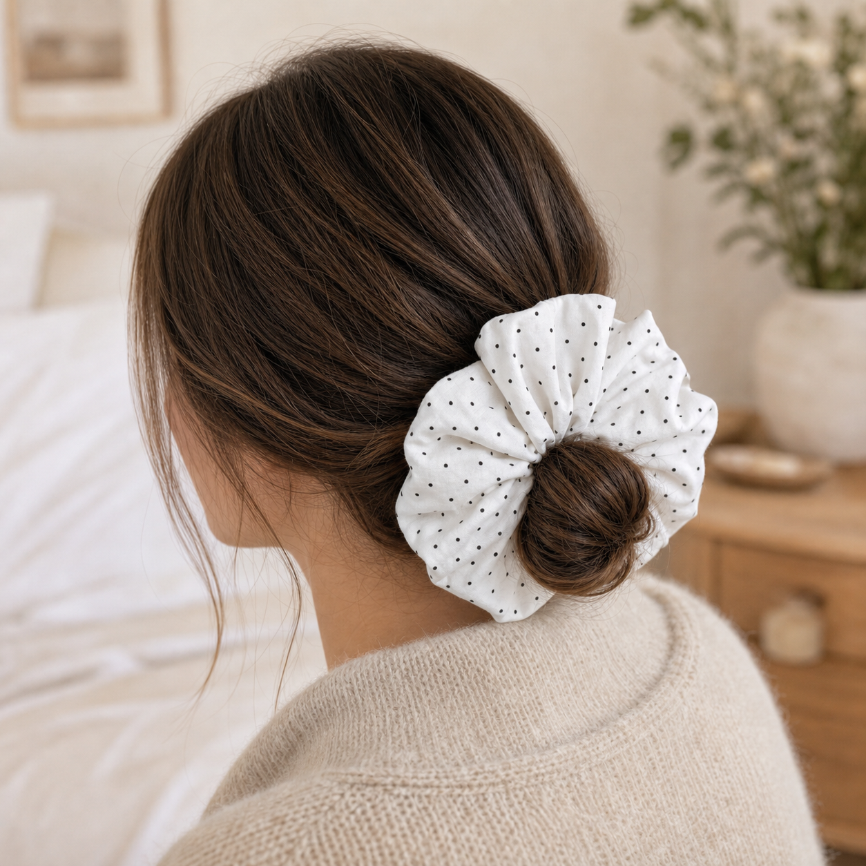 White black polka dot scrunchie styled in a low bun, rear view, cream knit sweater. Soft-focus bedroom background with white bedding, wooden nightstand, and vase of greenery with white flowers.