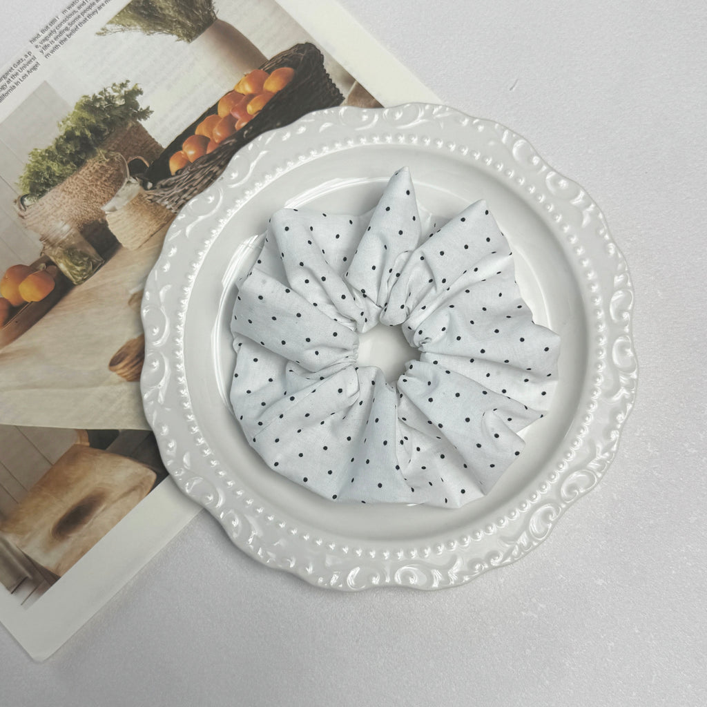 White black polka dot scrunchie placed on a glossy white ceramic plate with embossed scalloped rim, resting on an open magazine page featuring fruit basket photography, grey textured surface.