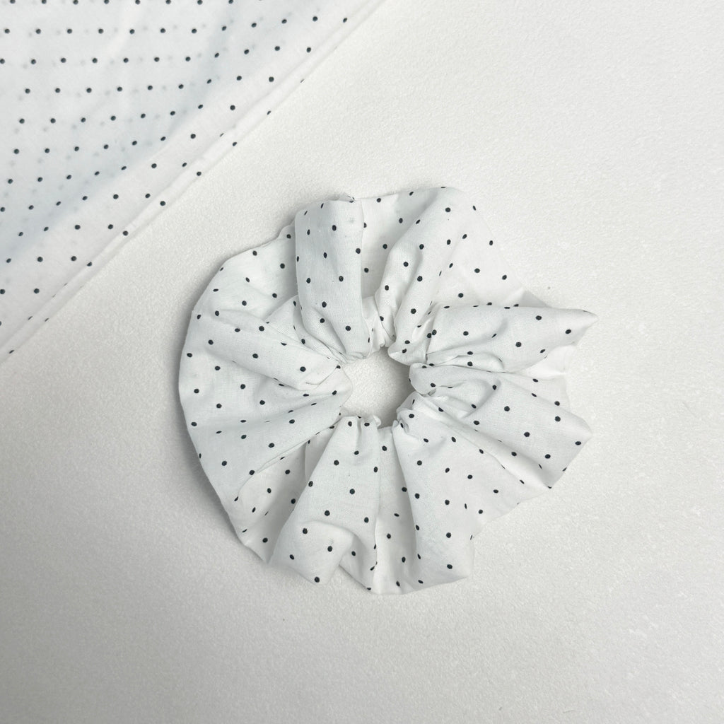 White black polka dot scrunchie displayed alongside a matching folded fabric swatch showing the full dot pattern, light grey textured surface.