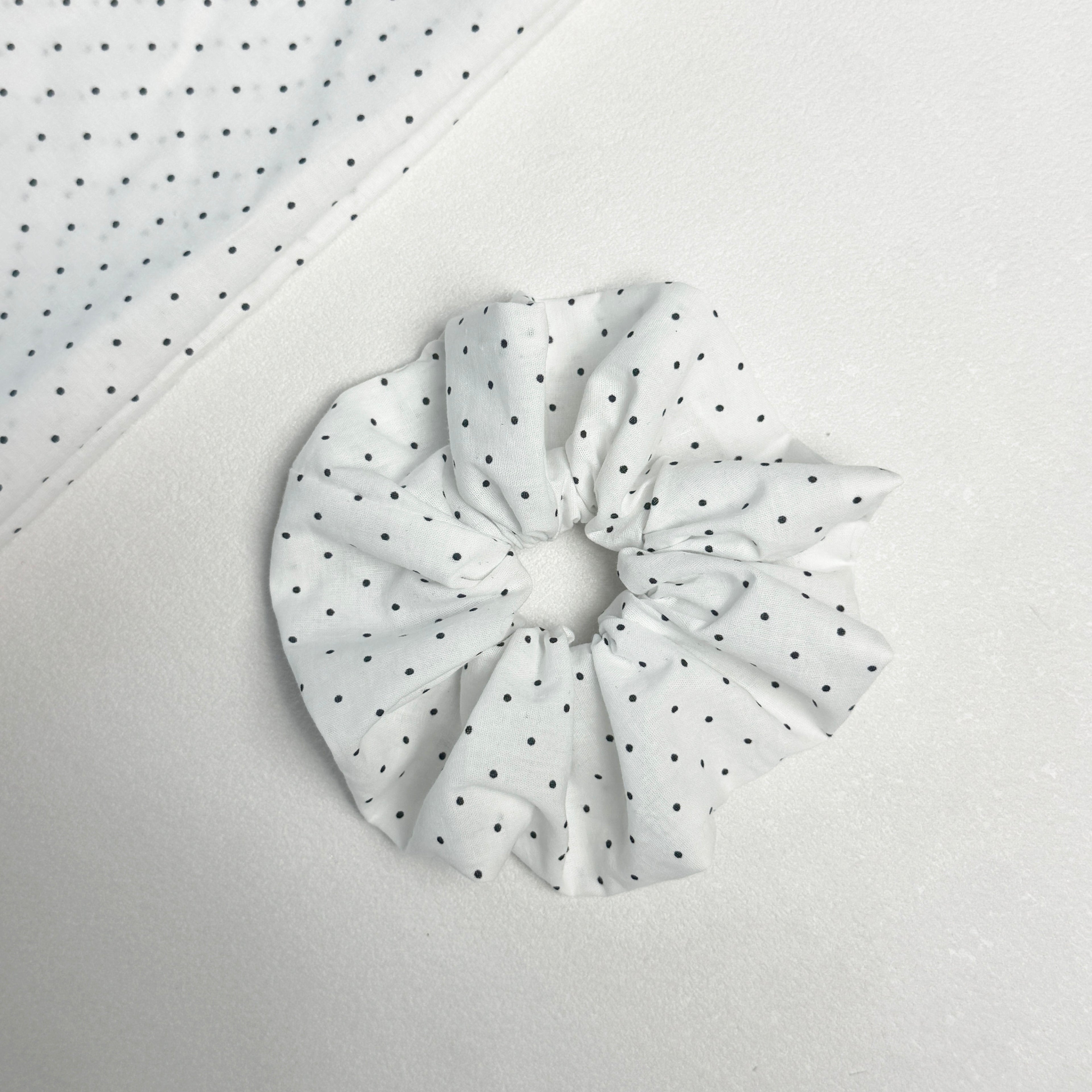 White black polka dot scrunchie displayed alongside a matching folded fabric swatch showing the full dot pattern, light grey textured surface.