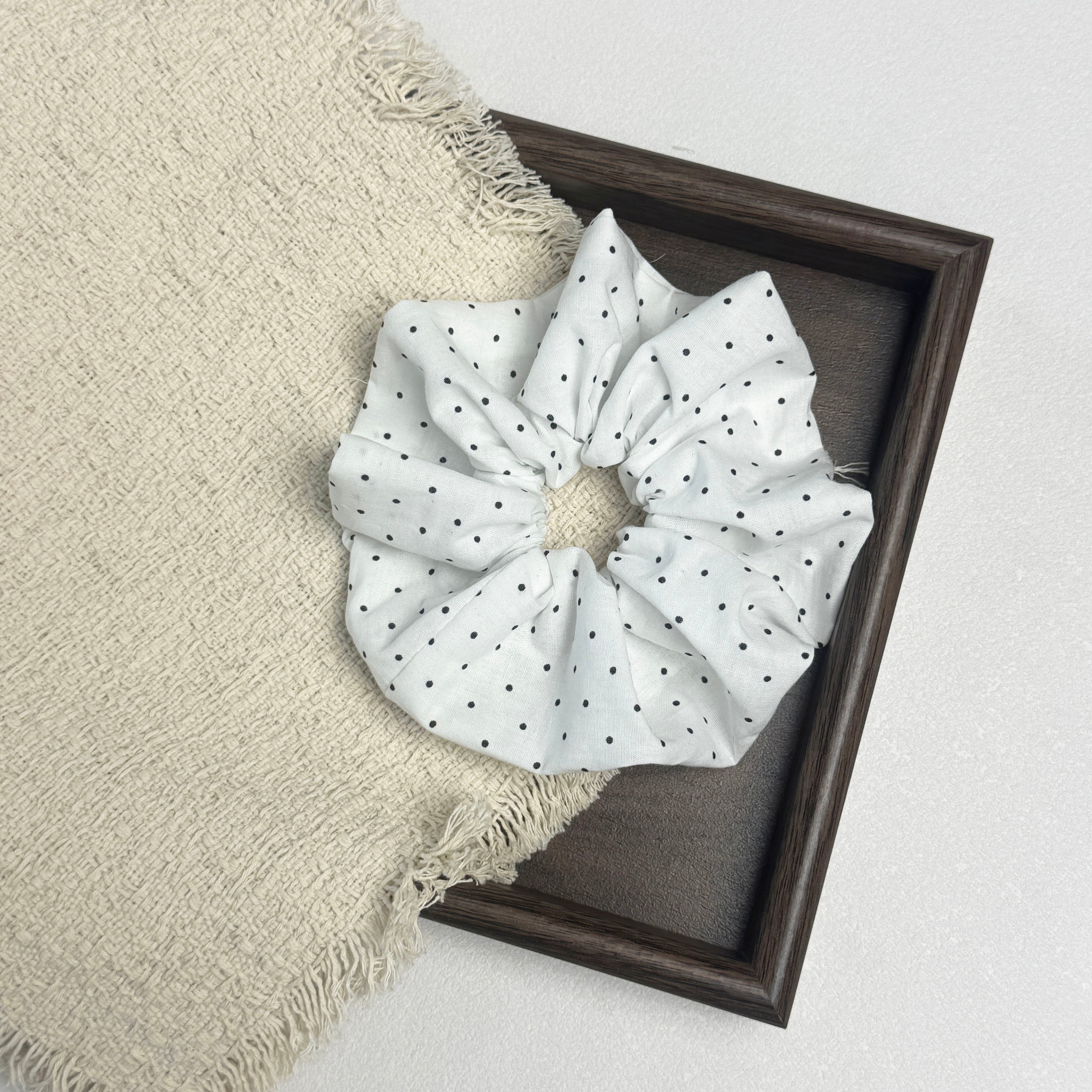 White black polka dot scrunchie flat-laid across a dark brown wooden picture frame and a frayed cream woven fabric with tasseled edges, white textured surface.
