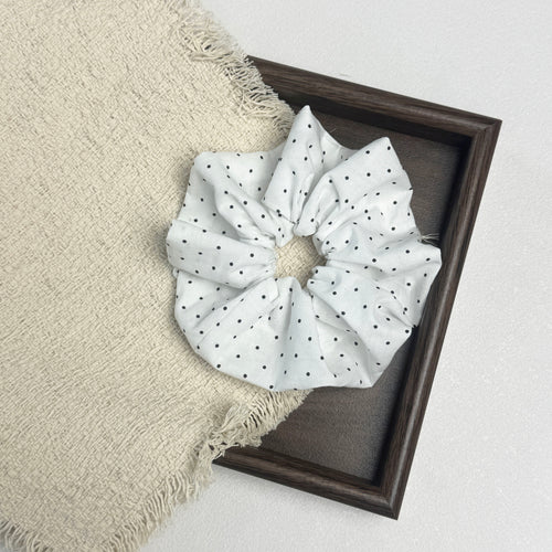 White black polka dot scrunchie flat-laid across a dark brown wooden picture frame and a frayed cream woven fabric with tasseled edges, white textured surface.