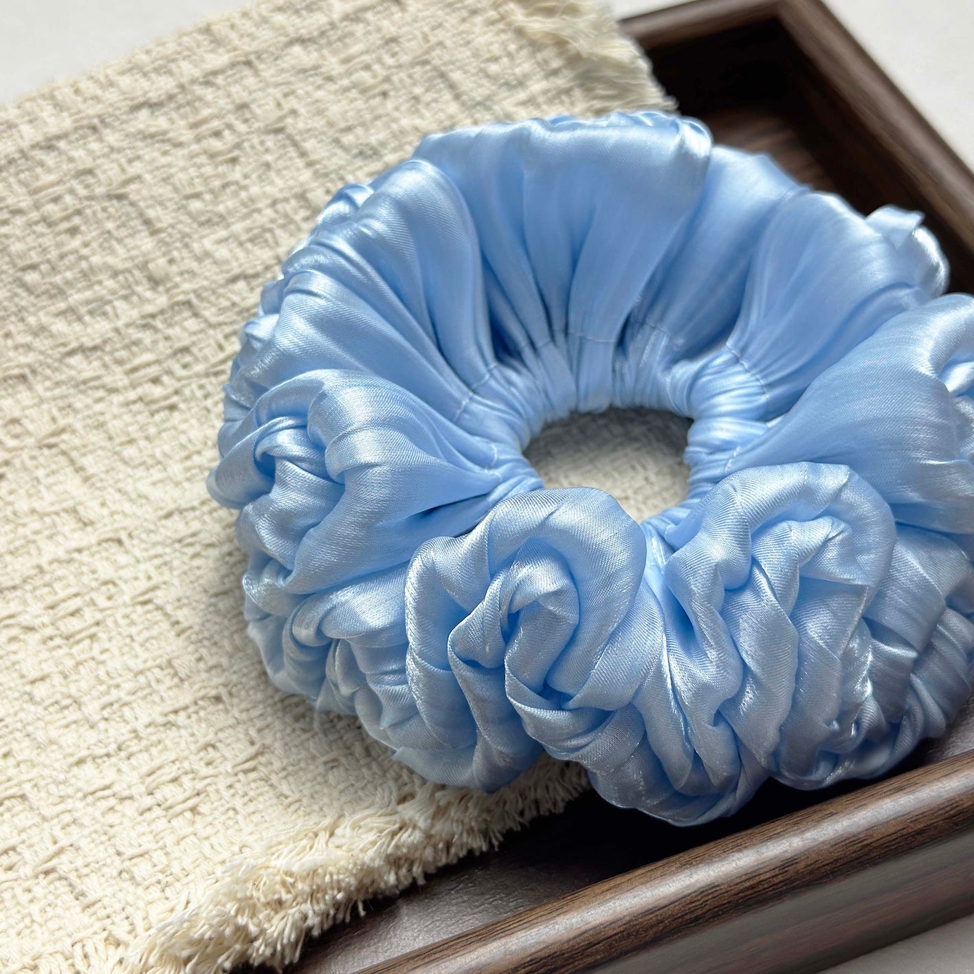Blue organza cloud scrunchie on a textured cream bouclé woven cloth inside a dark brown wooden tray with visible grain, soft natural lighting.