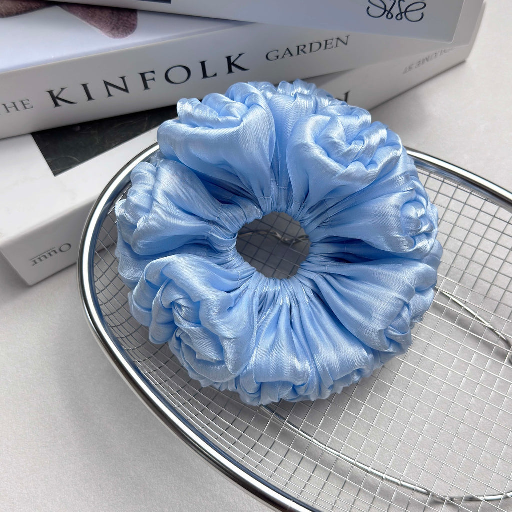 Blue organza cloud scrunchie resting in a polished silver wire mesh basket, placed on stacked white books, light grey surface.