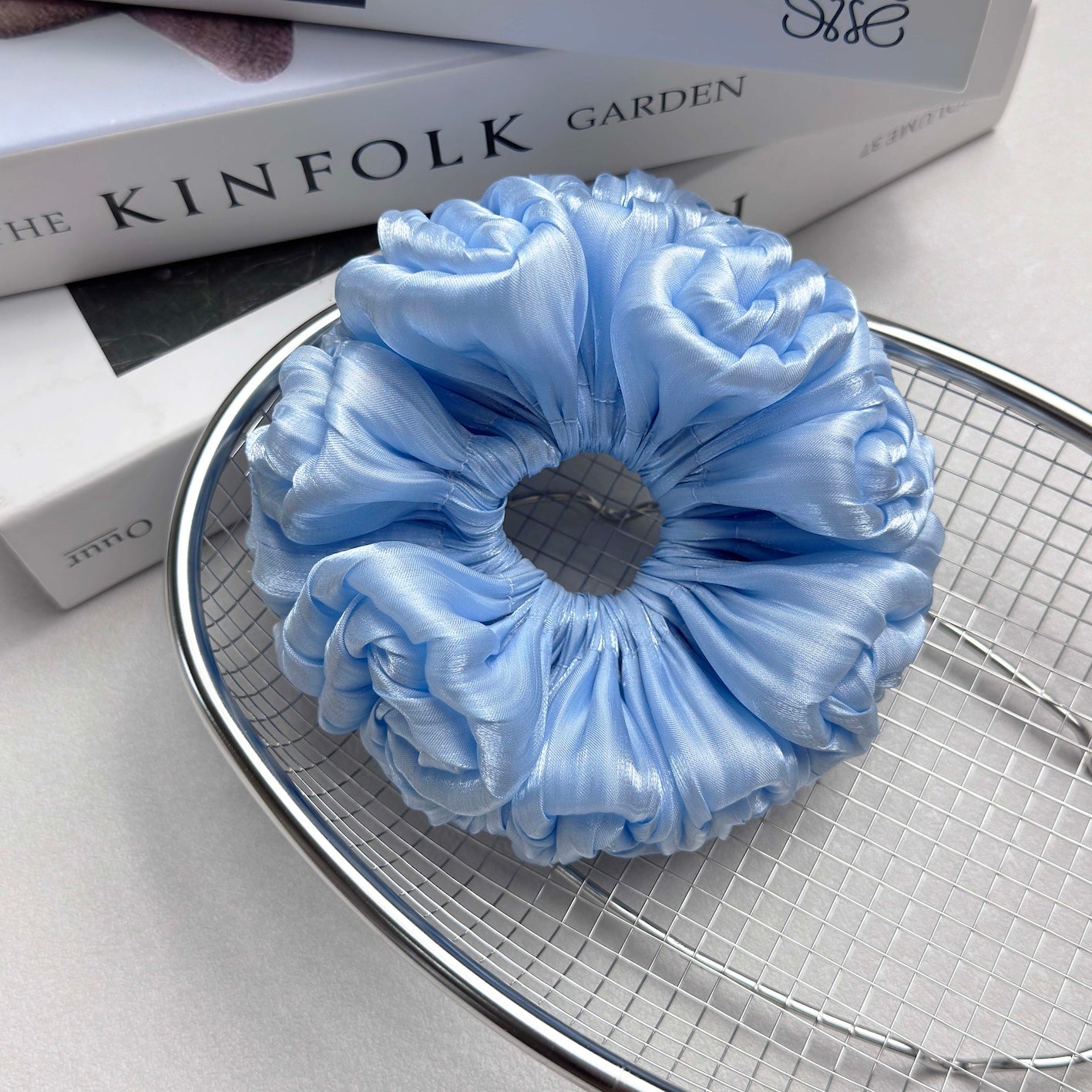 Blue organza cloud scrunchie resting in a polished silver wire mesh basket, placed on stacked white books, light grey surface.