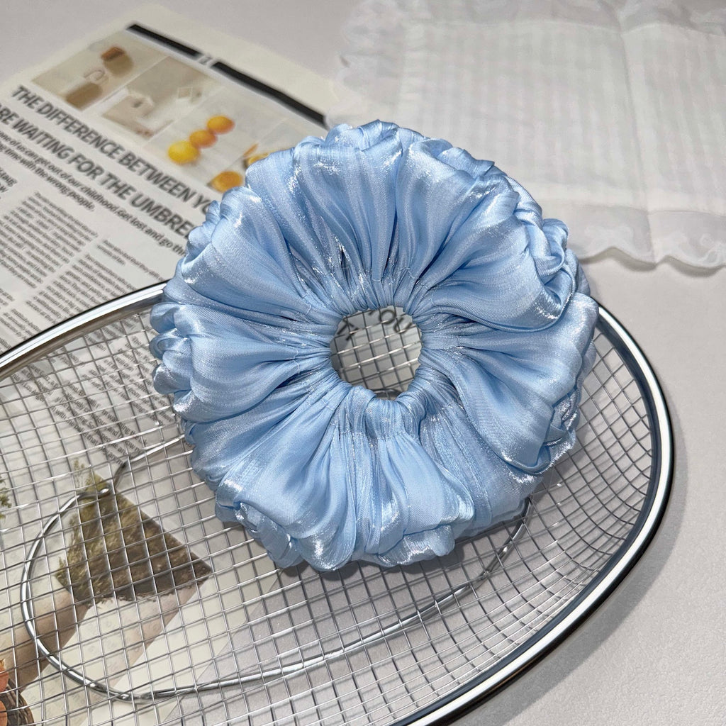 Blue organza cloud scrunchie resting on a silver wire mesh tray, placed over a magazine page with bold text and a white cloth with lace trim, grey-white surface.