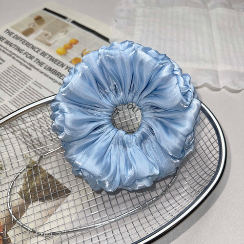 Blue organza cloud scrunchie resting on a silver wire mesh tray, placed over a magazine page with bold text and a white cloth with lace trim, grey-white surface.