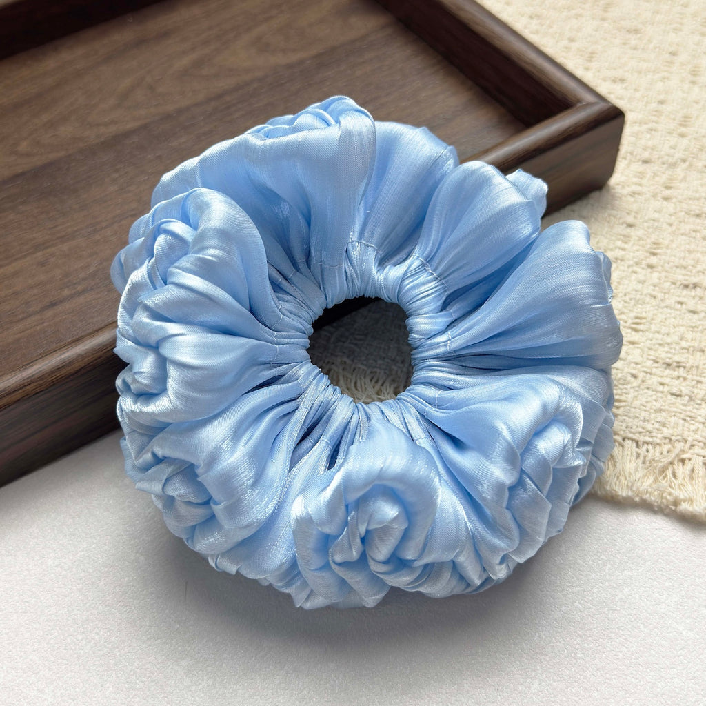 Blue organza cloud scrunchie on a grey-white surface with a dark brown wooden tray corner in the background and a fringed cream woven fabric to the right, flat-lay composition.