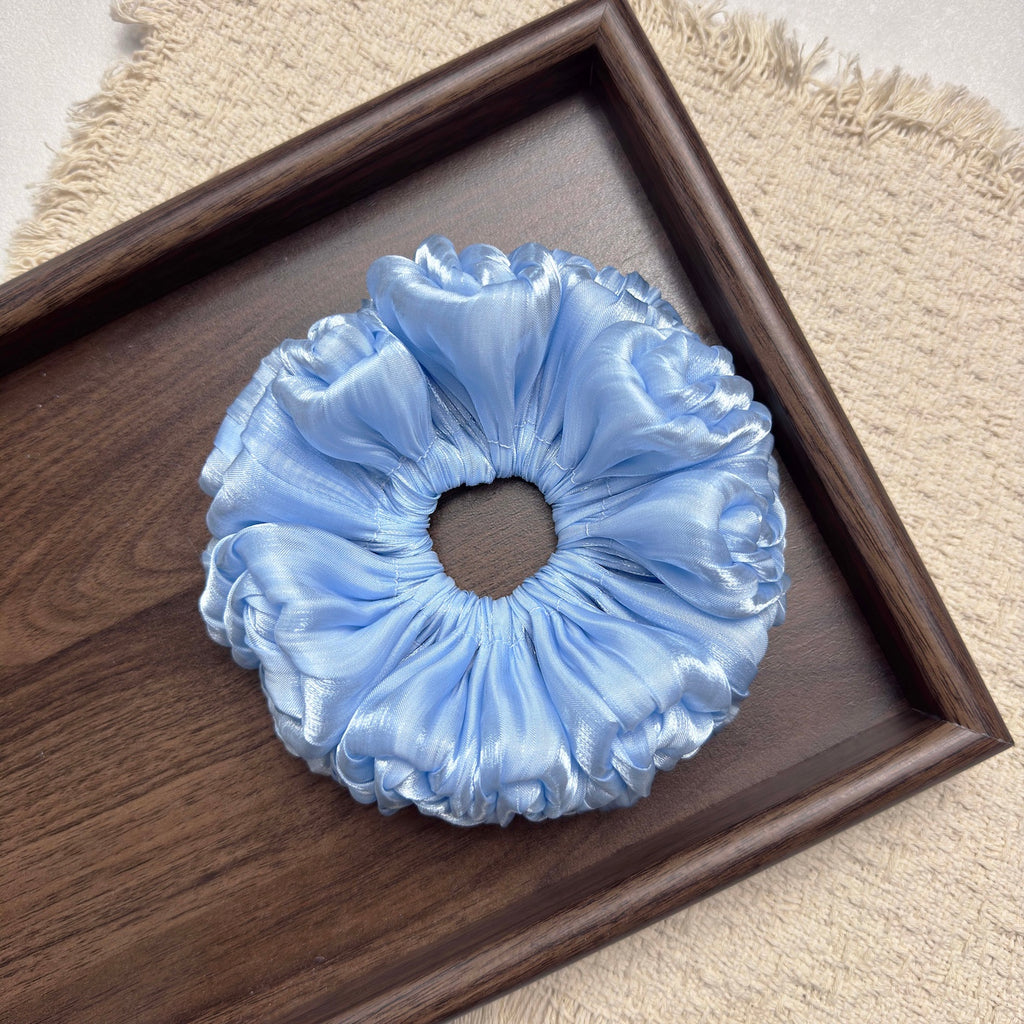Blue organza cloud scrunchie centered in a dark brown wooden tray on a fringed cream woven textile, flat-lay composition.