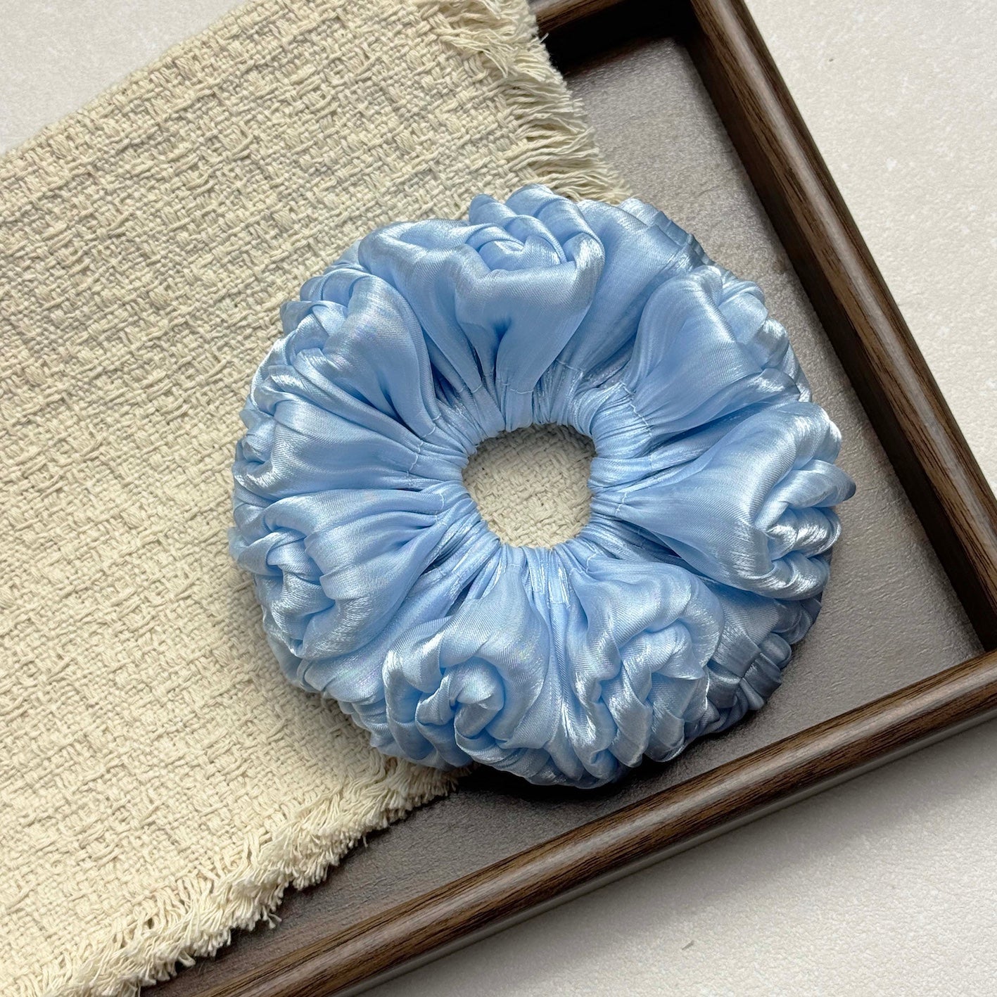 Blue organza cloud scrunchie resting on a fringed cream waffle-weave cloth inside a dark brown wooden tray with grey interior, flat-lay composition.