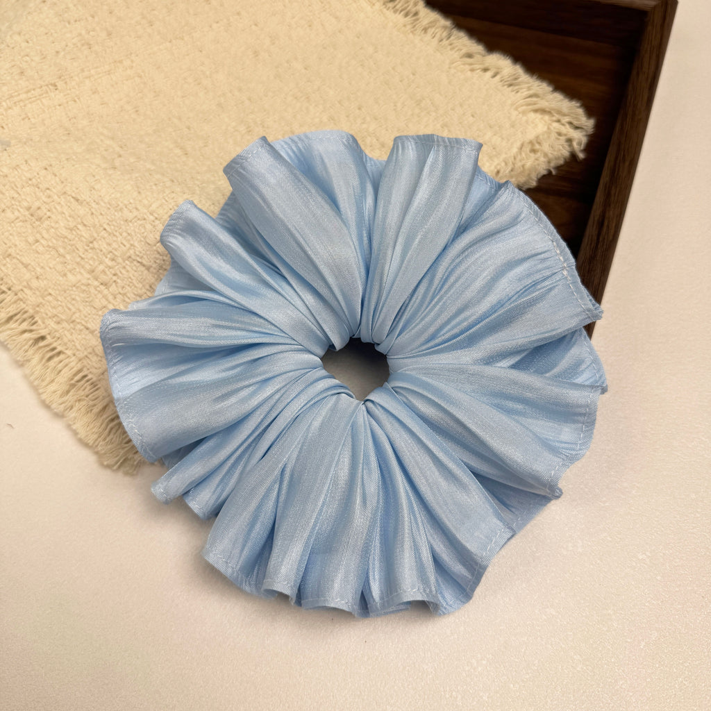 Soft blue pleated satin scrunchie on a white surface beside a frayed cream burlap-textured fabric and the corner of a dark brown wooden tray, flat-lay composition.