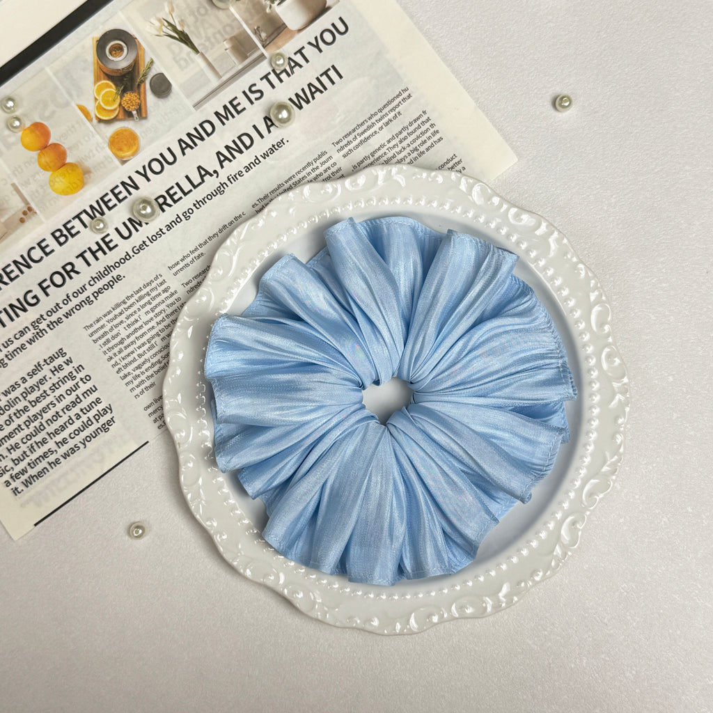 Soft blue pleated satin scrunchie placed on a white ceramic plate with embossed scalloped rim, resting on a magazine page with citrus photography and text, scattered white faux pearls, grey-white surface.