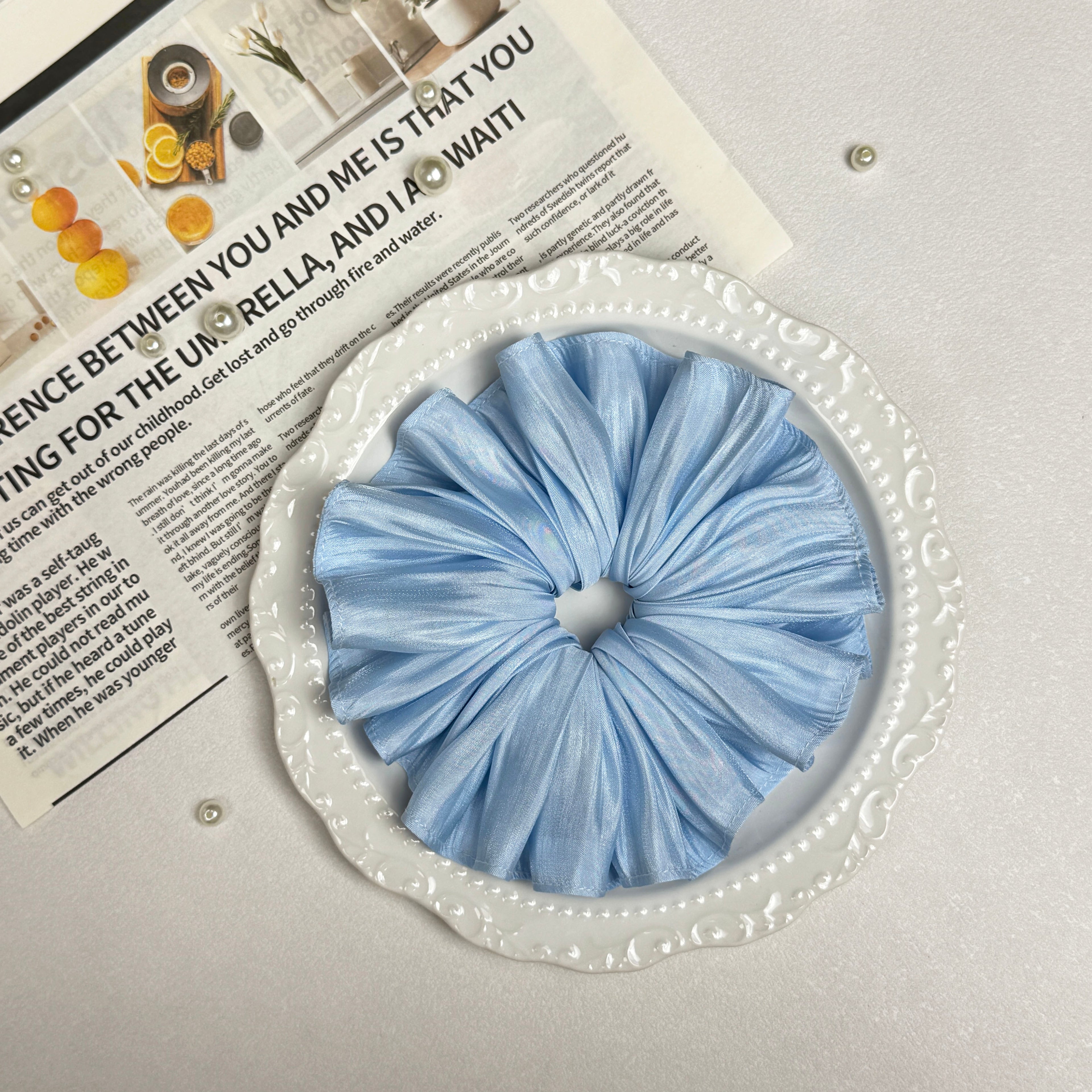 Soft blue pleated satin scrunchie placed on a white ceramic plate with embossed scalloped rim, resting on a magazine page with citrus photography and text, scattered white faux pearls, grey-white surface.