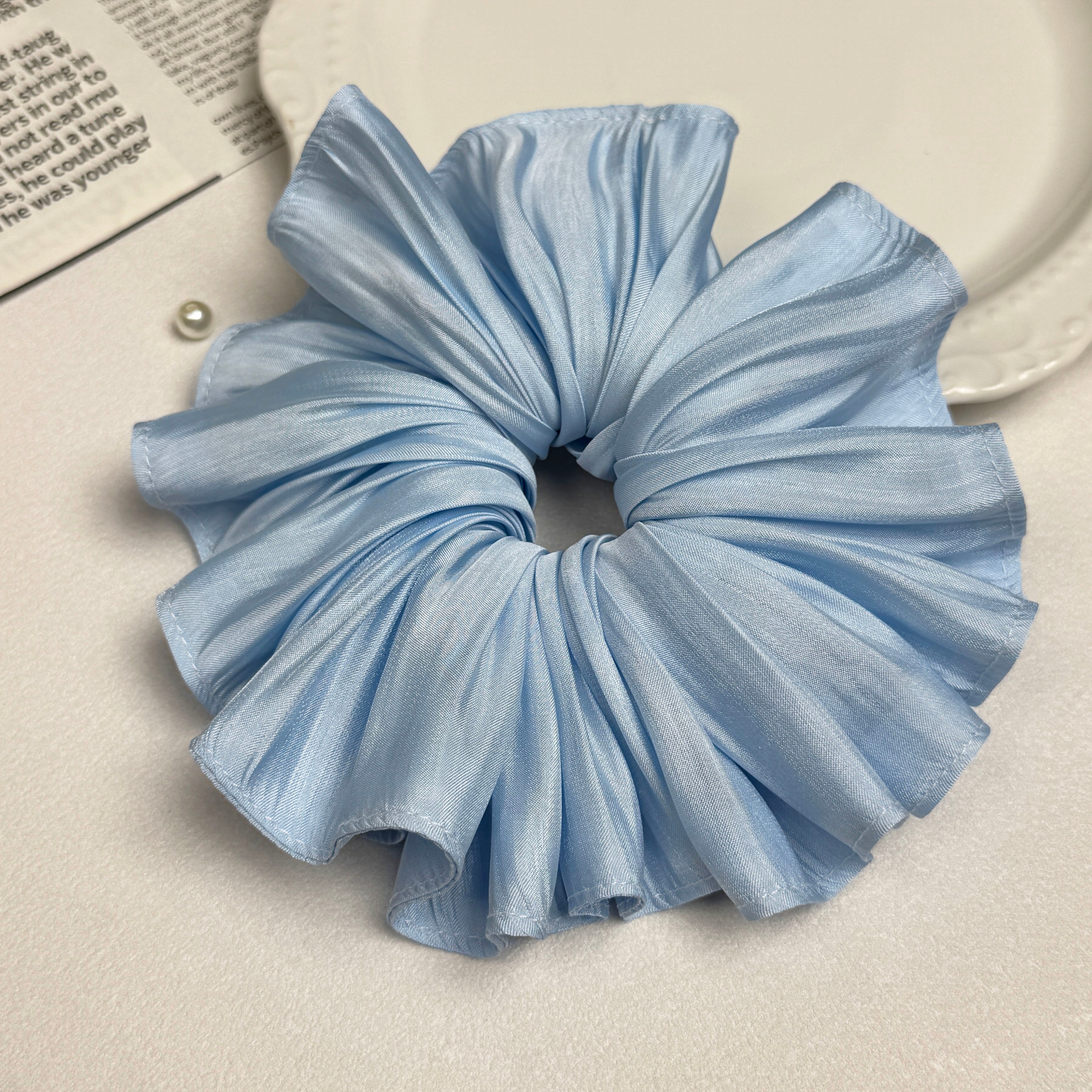 Soft blue pleated satin scrunchie alongside a white ceramic plate with beaded scalloped rim, an open magazine page with printed text, and a single white faux pearl, clean white surface, flat-lay composition.