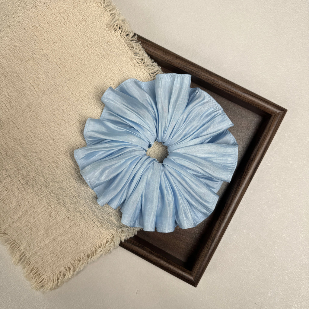 Soft blue pleated satin scrunchie flat-laid across a dark brown wooden tray and a frayed cream woven fabric with tasseled edges, grey-white textured surface.