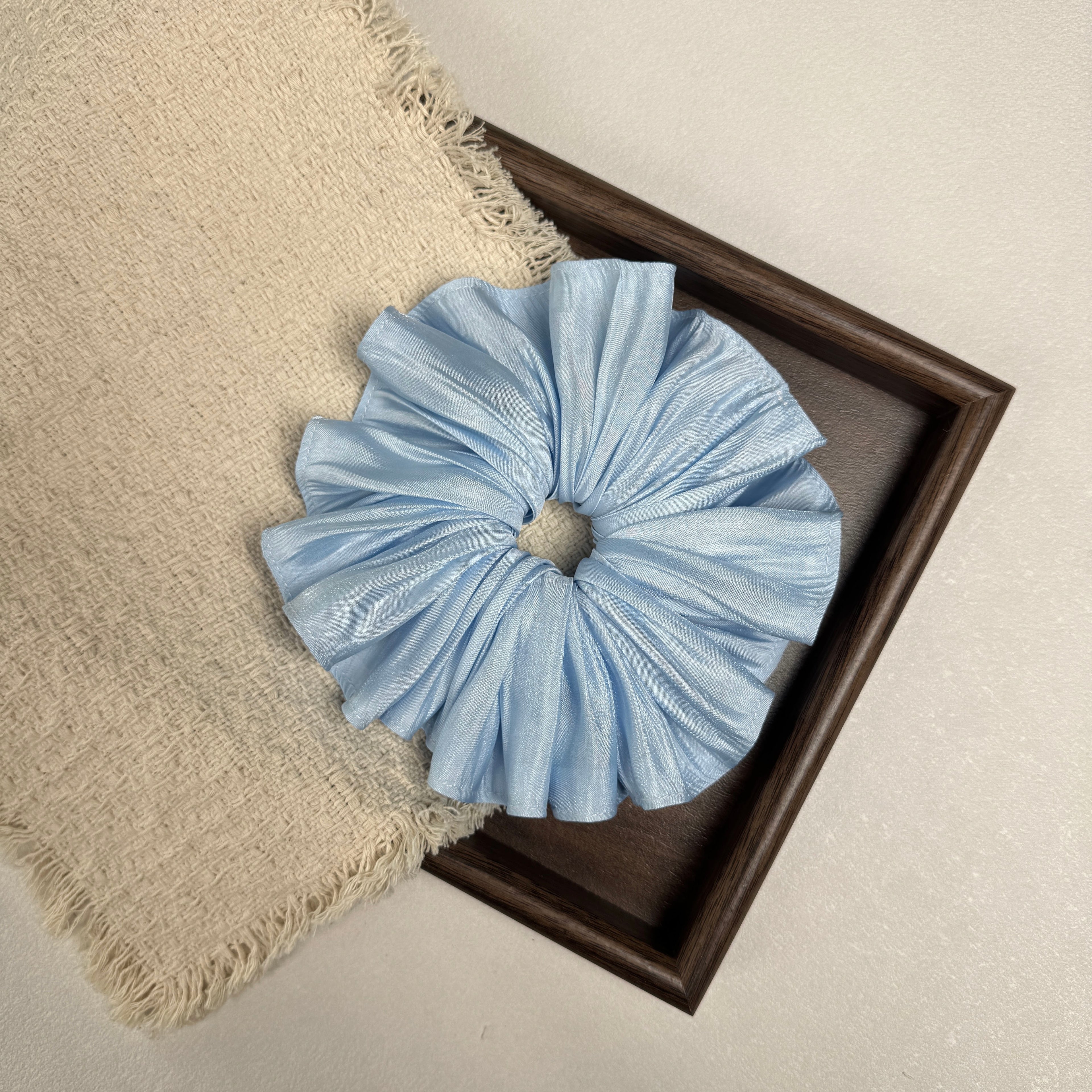 Soft blue pleated satin scrunchie flat-laid across a dark brown wooden tray and a frayed cream woven fabric with tasseled edges, grey-white textured surface.