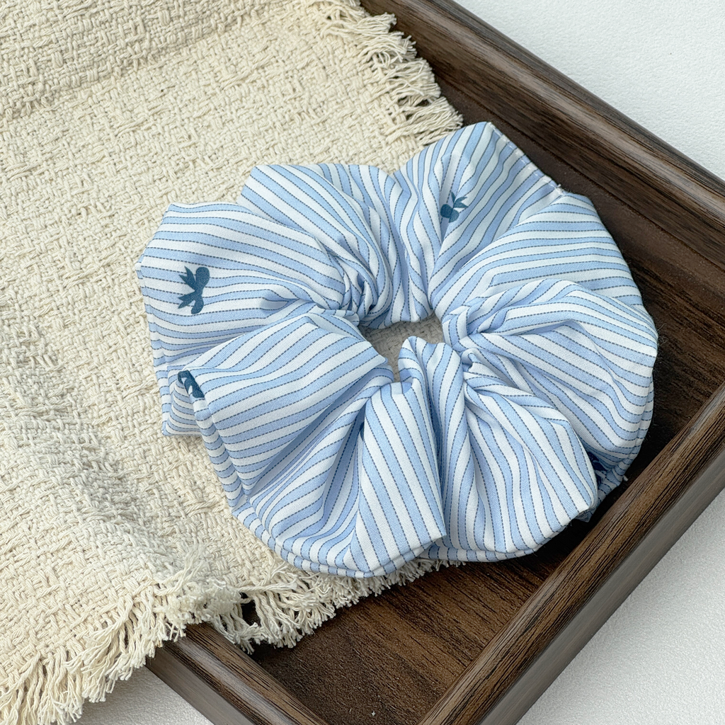 Close-up detail of a blue striped bow scrunchie on a wooden tray, showcasing linen texture and no-crease design.