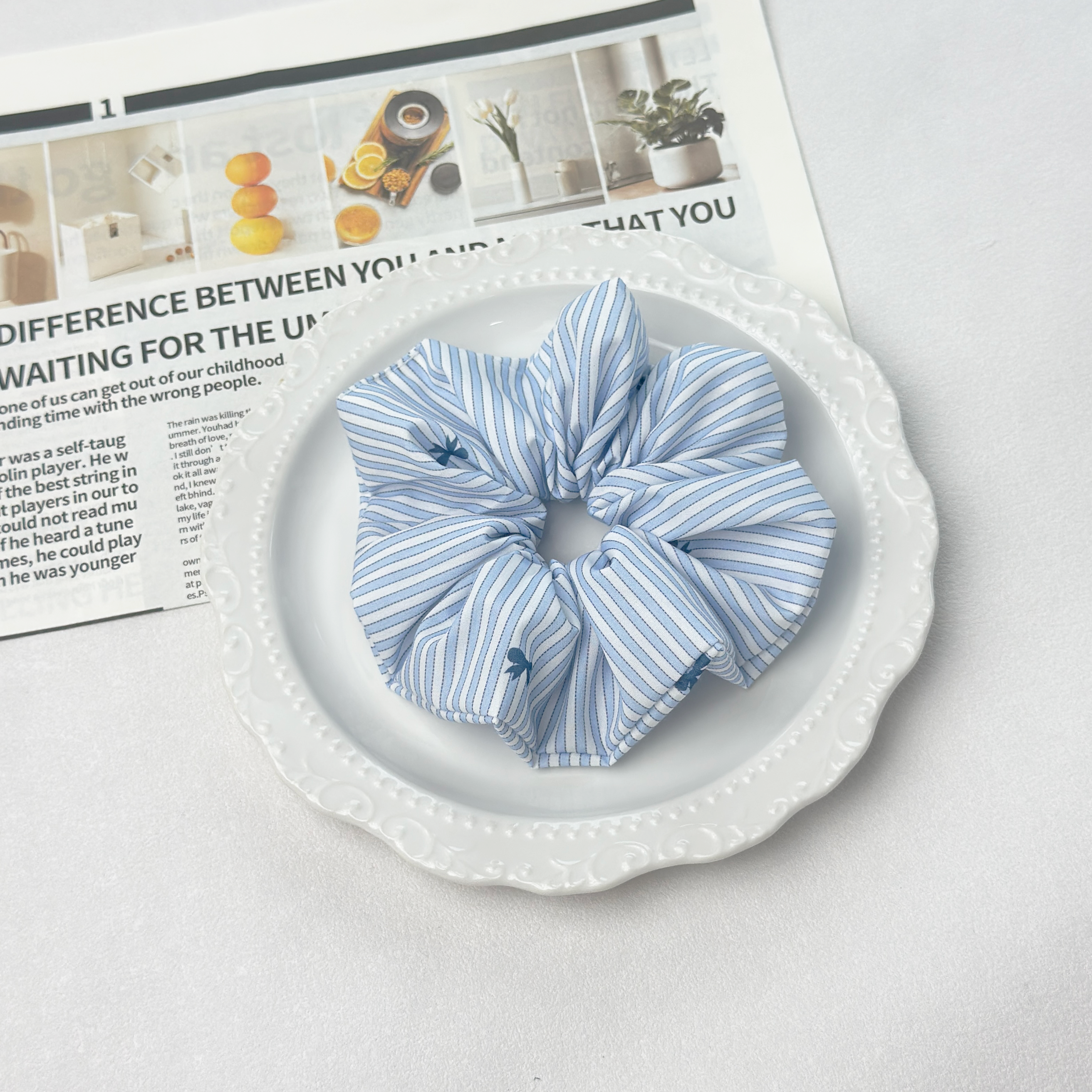 Top-down view of blue striped bow scrunchie on a white decorative plate, stylish preppy hair care.