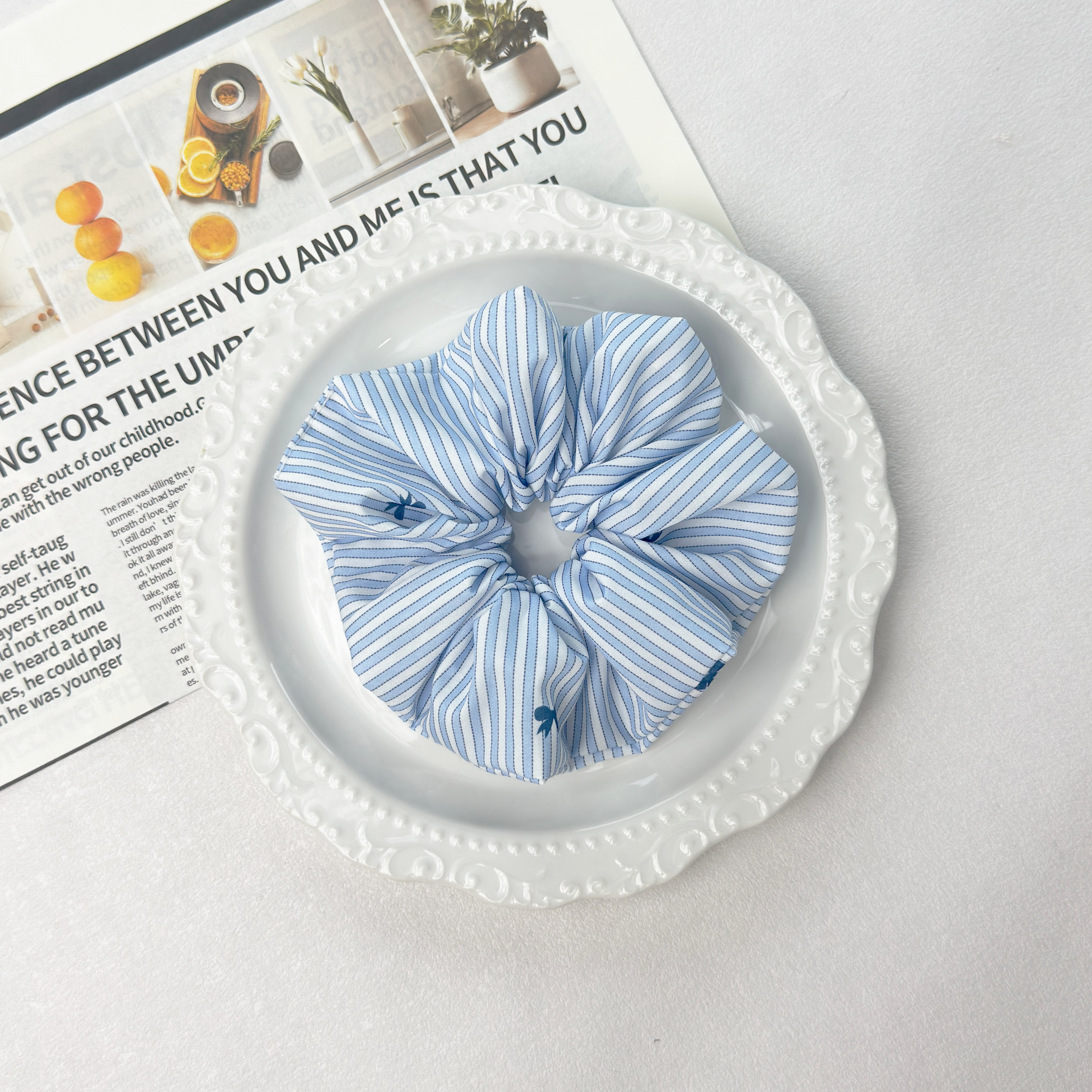 Blue striped bow scrunchie on a white wavy decorative plate with a newspaper background, preppy linen blend.