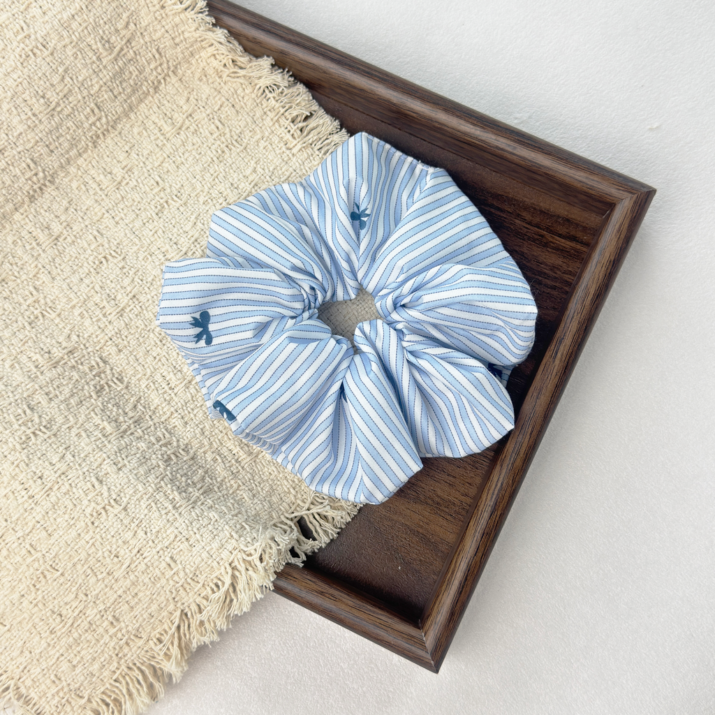 Blue striped bow scrunchie on a wooden tray with beige fringed woven cloth, premium linen blend hair tie.