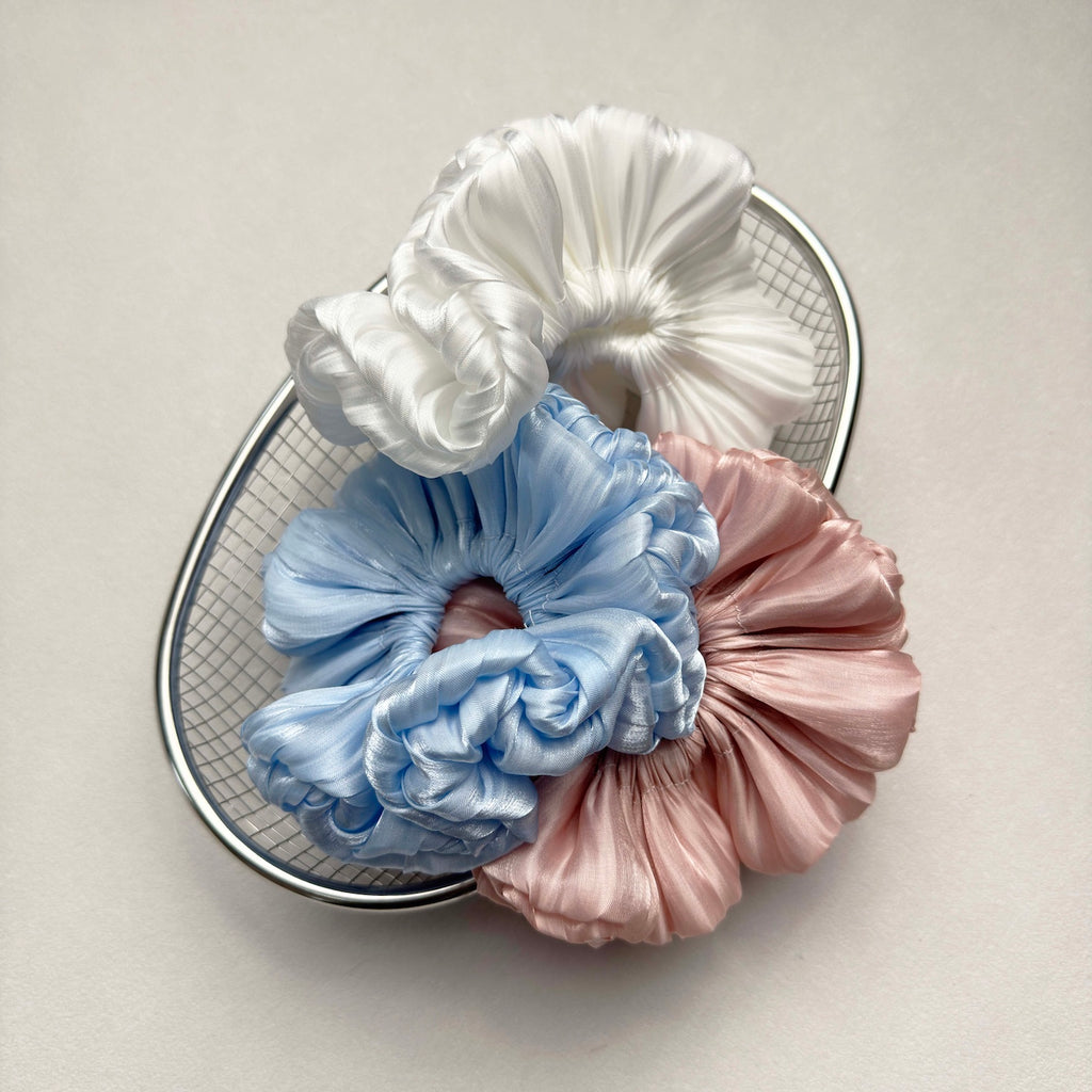 Three organza cloud scrunchies in white, blue, and pink clustered together inside a silver wire mesh basket, grey-white pebbled surface.