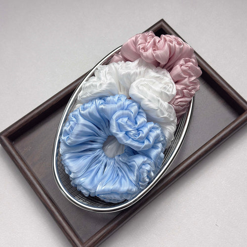 Three organza cloud scrunchies in pink, white, and blue stacked inside a silver wire mesh basket, placed within a dark brown wooden tray, grey-white surface.