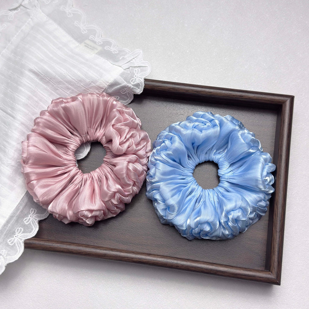 Two organza cloud scrunchies in dusty pink and powder blue inside a dark brown wooden tray, with a piece of white striped lace-trimmed fabric draped over the top left corner, grey-white surface.