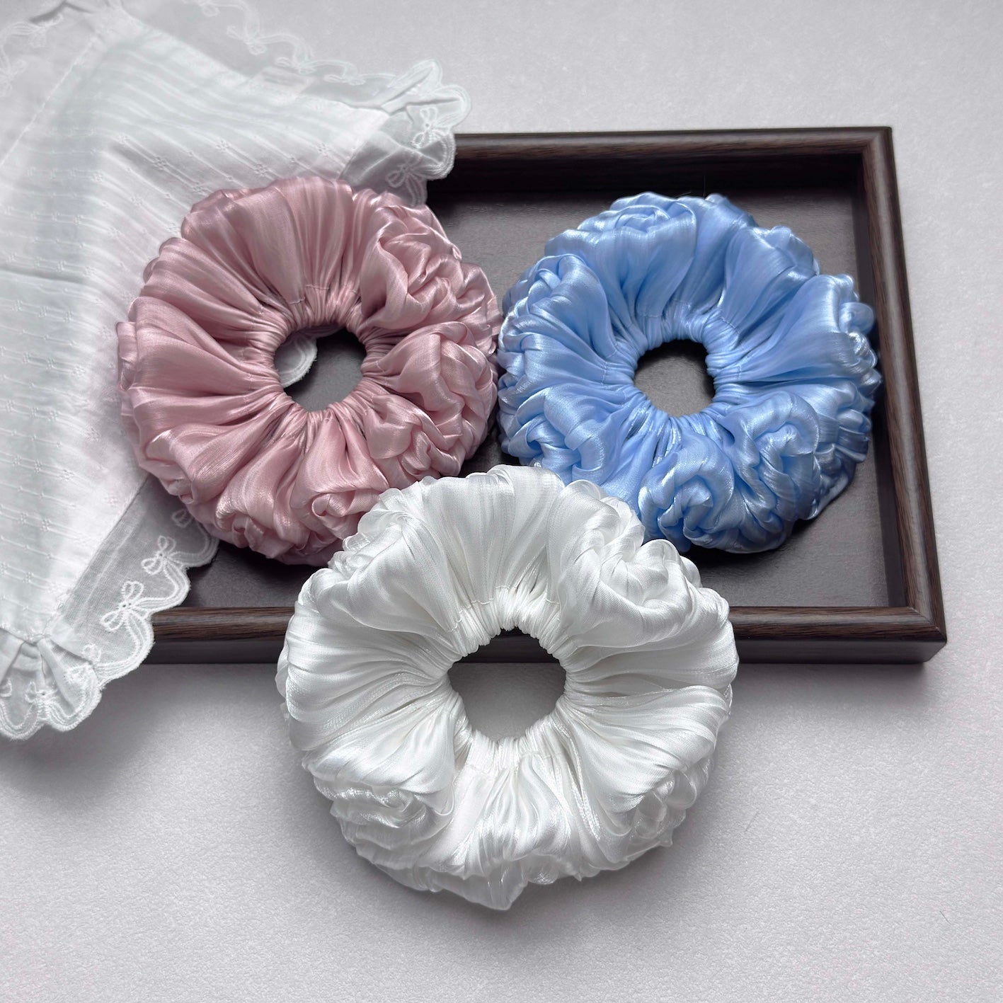 Three organza cloud scrunchies in pink, blue, and white arranged around a dark brown wooden frame, with a piece of white scalloped lace fabric in the top left corner, grey-white textured surface.
