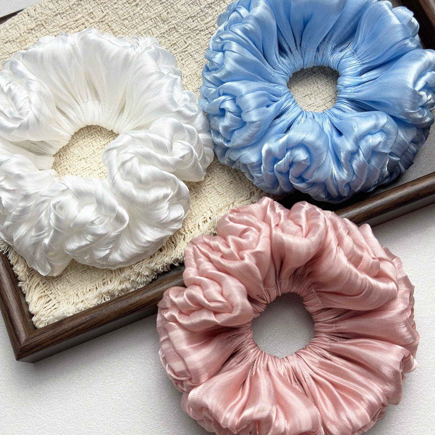 Three organza cloud scrunchies in white, blue, and pink on a cream burlap-textured fabric with frayed edges inside a dark brown wooden tray, white pebbled surface.