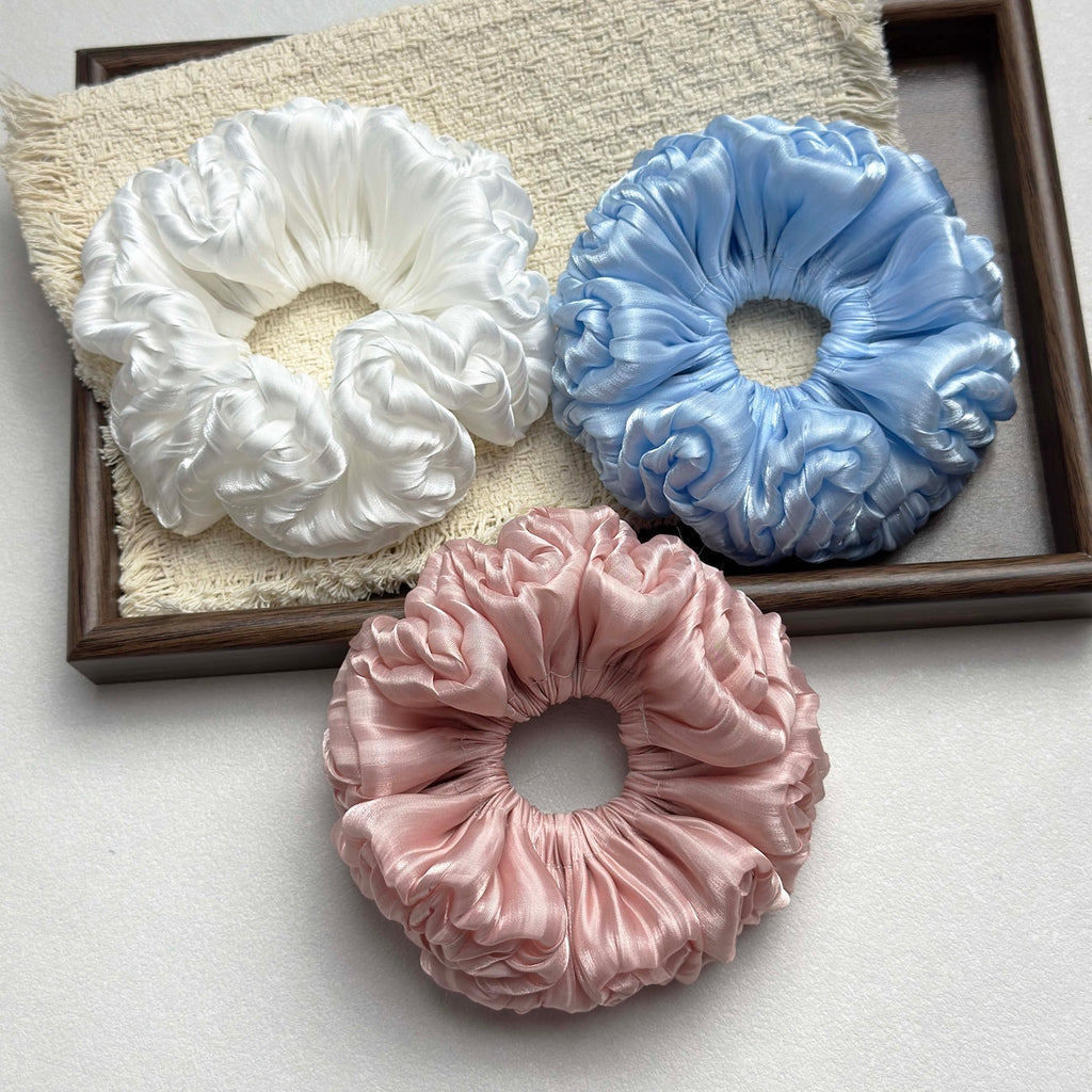 Three organza cloud scrunchies in white, blue, and pink arranged on a fringed cream woven fabric inside a dark brown wooden tray, flat-lay composition.