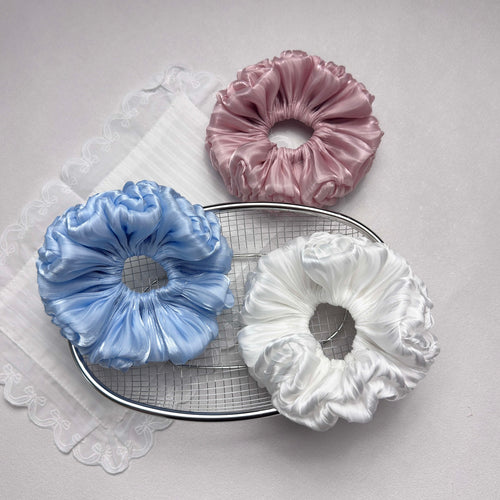 Three organza cloud scrunchies in blue, pink, and white arranged around a silver wire mesh tray with a white scalloped lace fabric, grey-white textured surface, flat-lay composition.