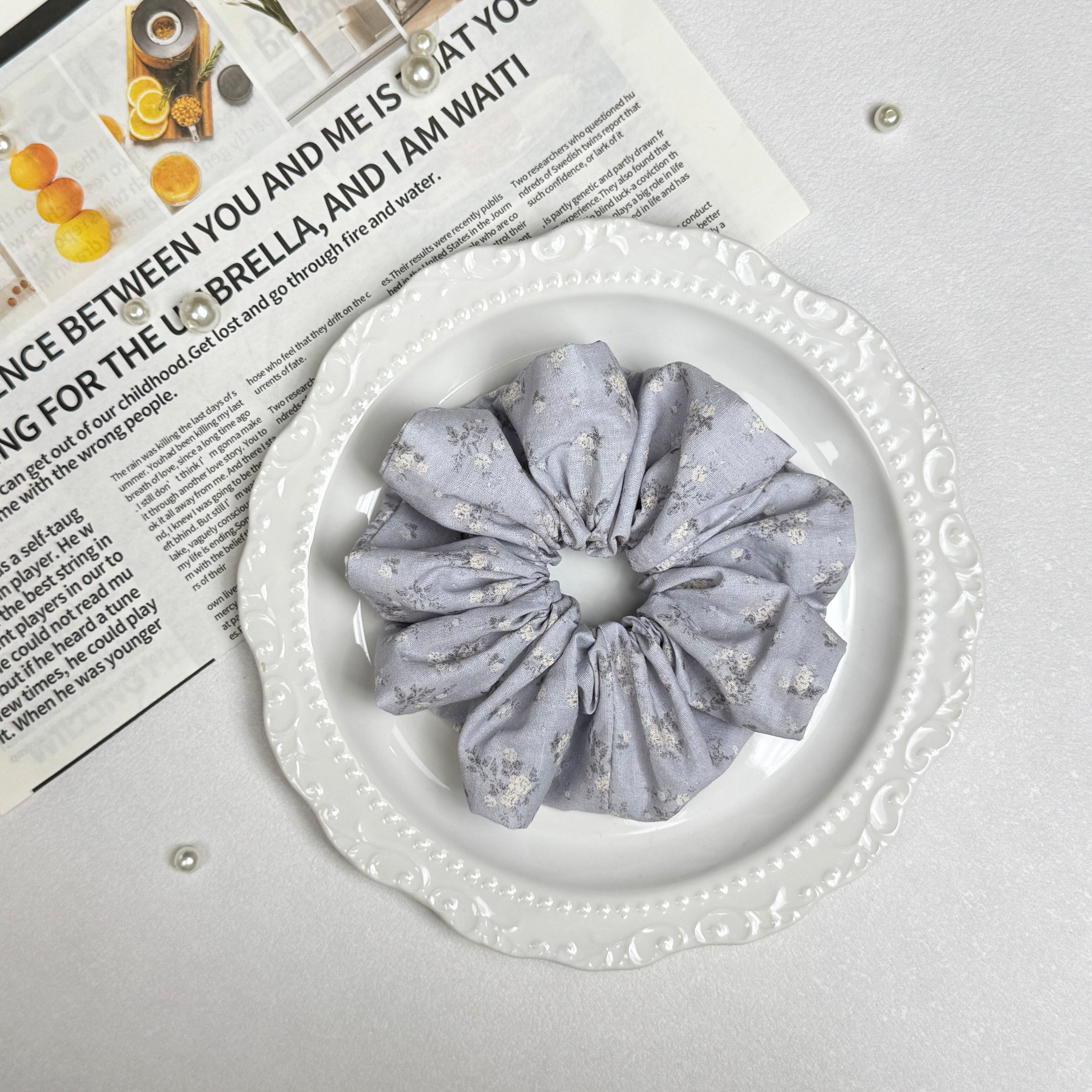 Dusty blue ditsy floral scrunchie placed on a white ceramic plate with embossed scalloped rim, resting on a magazine page with citrus photography and text, scattered white faux pearls, grey-white surface.