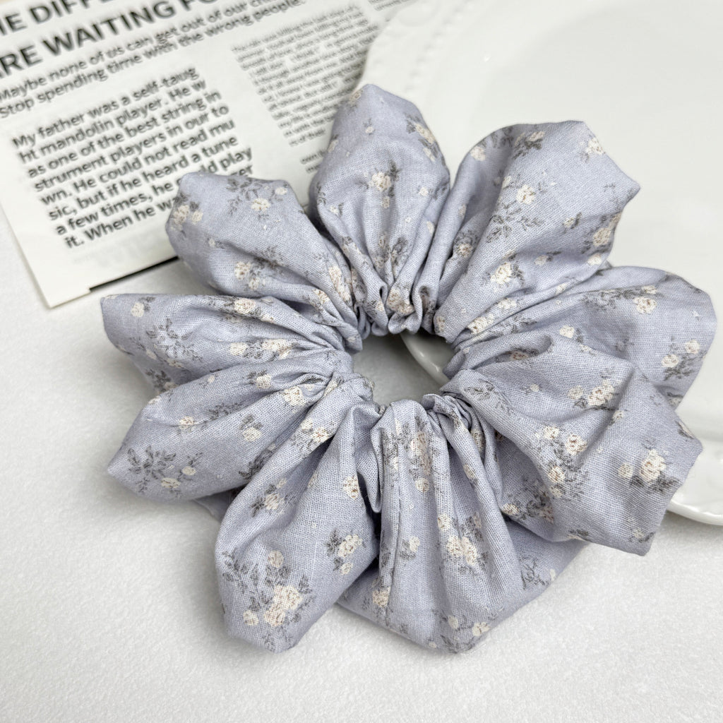 Dusty blue ditsy floral scrunchie alongside a white ceramic plate with beaded scalloped rim and a magazine page with printed text, clean white surface, flat-lay composition.