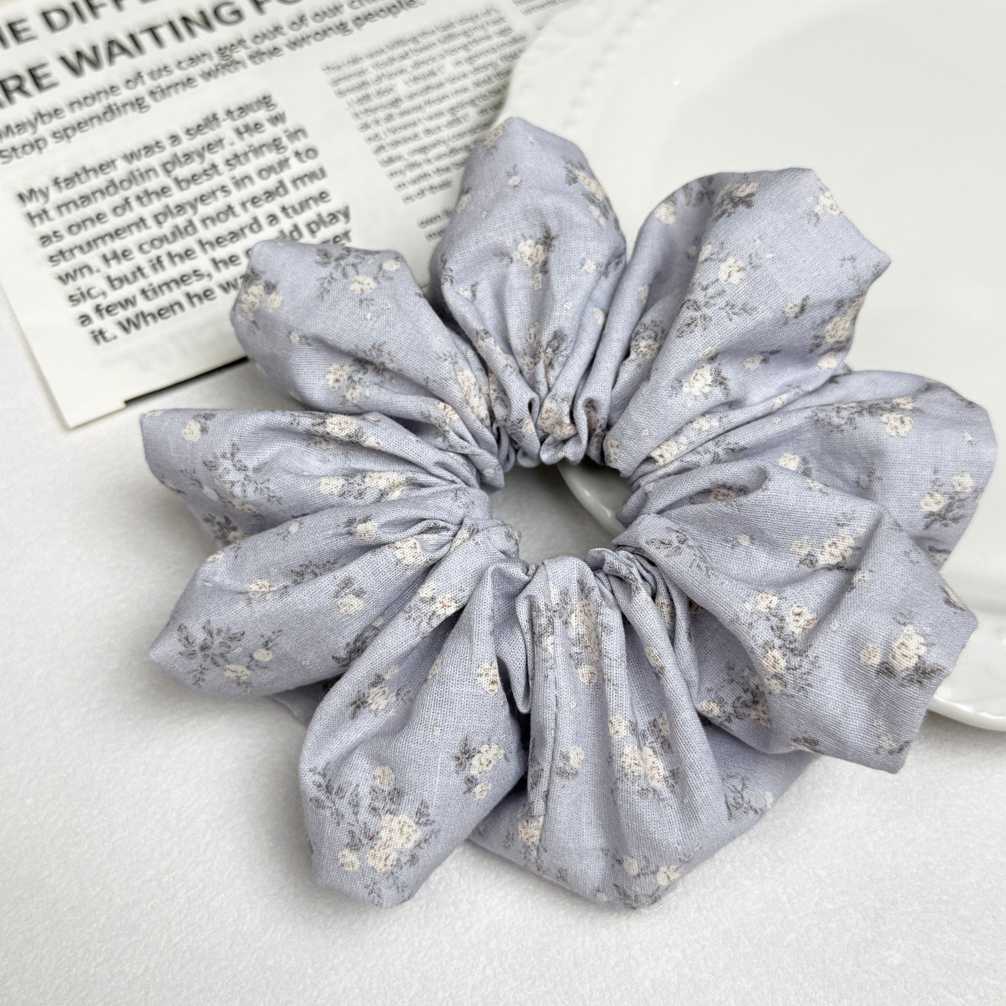 Dusty blue ditsy floral scrunchie alongside a white ceramic plate with beaded scalloped rim and a magazine page with printed text, clean white surface, flat-lay composition.