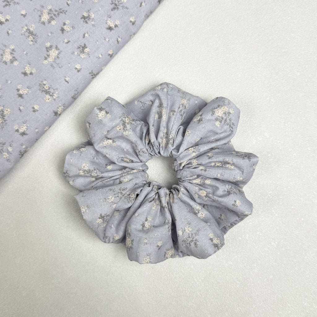 Dusty blue ditsy floral scrunchie displayed alongside a matching triangular fabric swatch showing the full white floral print, white pebbled texture surface.