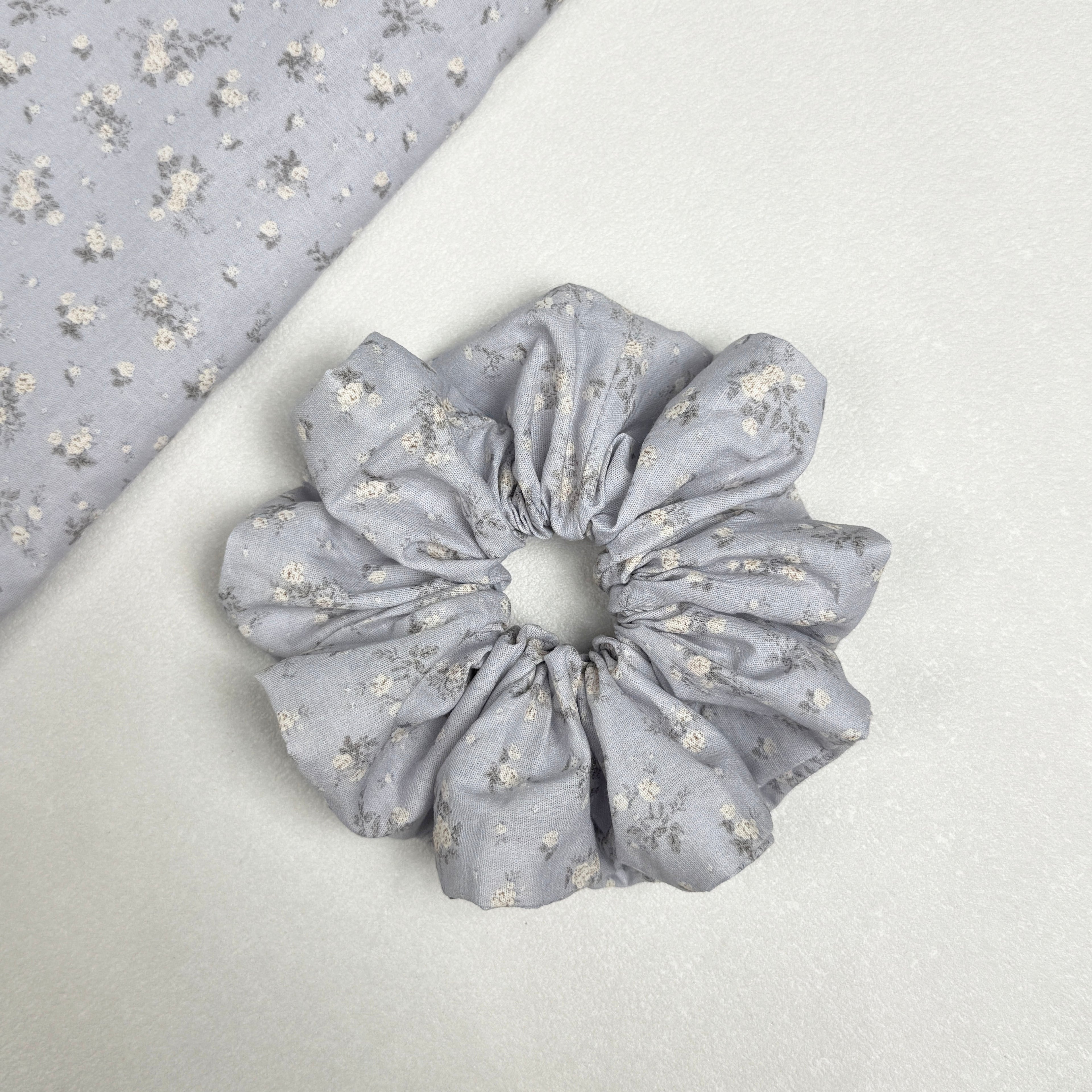 Dusty blue ditsy floral scrunchie displayed alongside a matching triangular fabric swatch showing the full white floral print, white pebbled texture surface.