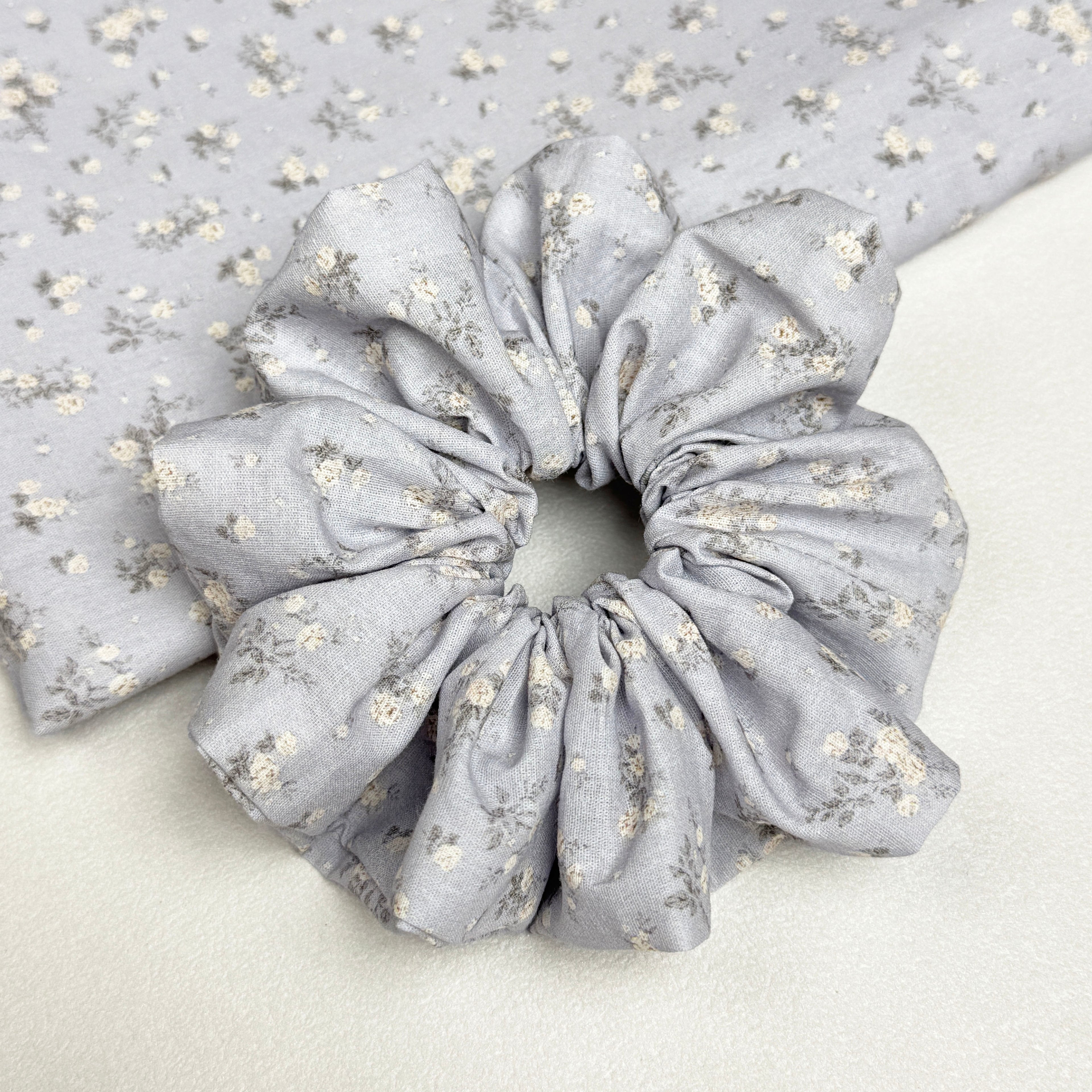 Dusty blue ditsy floral scrunchie displayed alongside a matching neatly folded fabric piece showing the cream blossom and grey stem pattern, white textured surface.