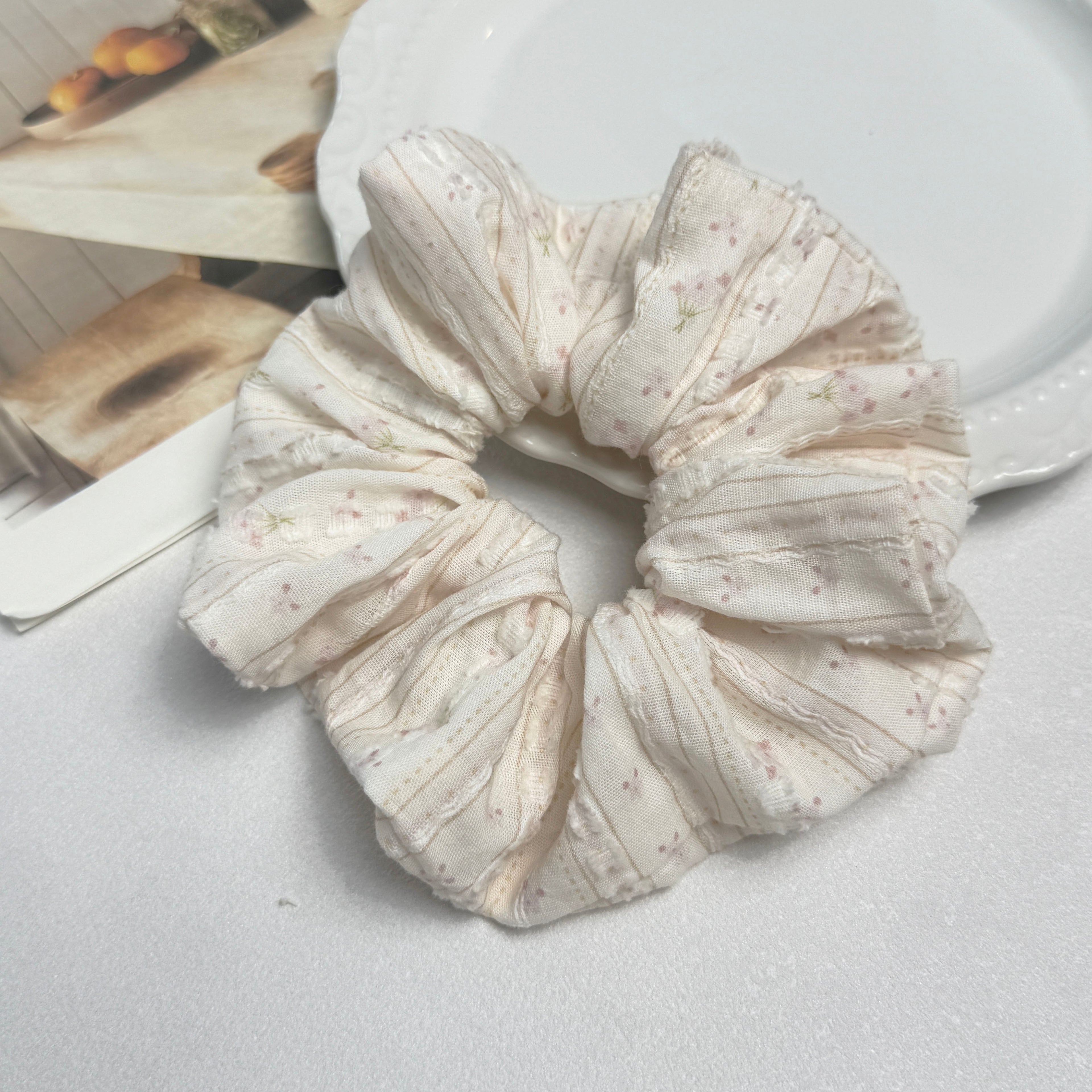 Cream floral bouquet print scrunchie alongside a white ceramic plate with beaded scalloped rim and a magazine page with warm-toned fruit photography, top-down flat lay, white textured surface.