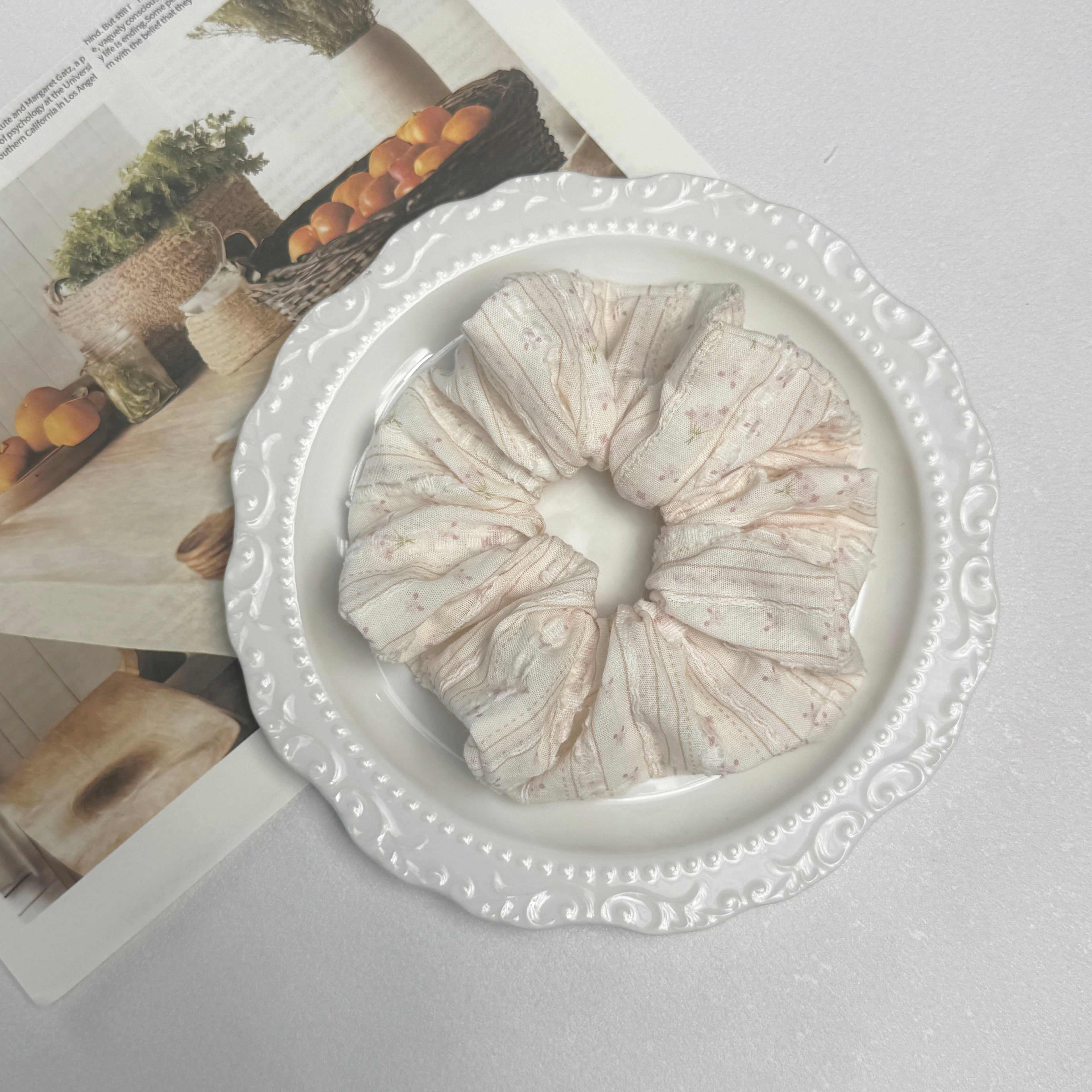 Cream floral bouquet print scrunchie placed on a glossy white ceramic plate with embossed scalloped rim, resting on a magazine page featuring rustic fruit basket photography, grey textured surface.