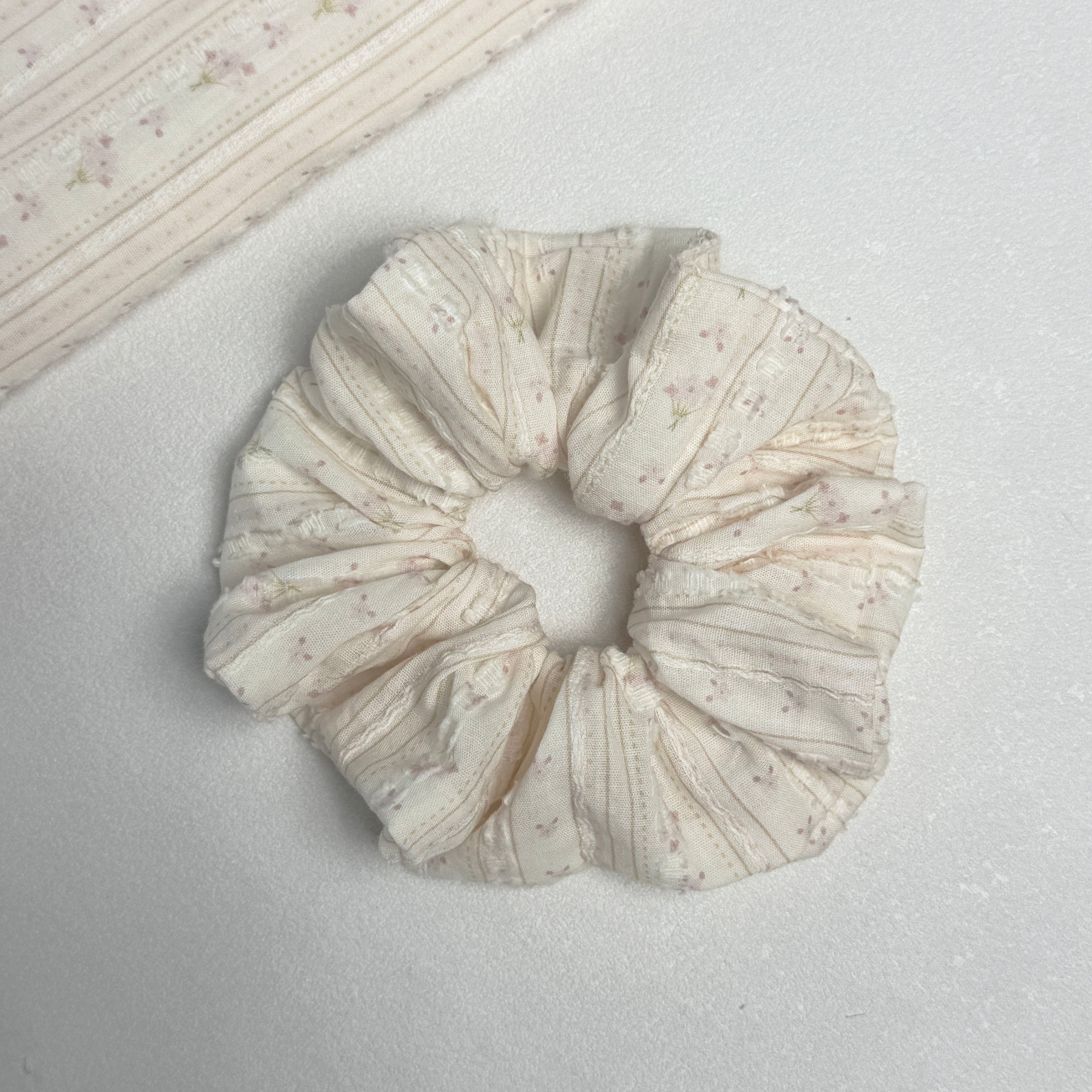 Cream floral bouquet print scrunchie displayed alongside a matching fabric swatch showing the full floral print, vertical ribbed texture, and raised tufted dots, white textured surface.