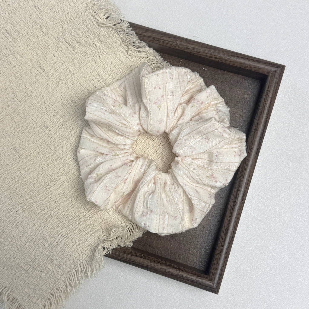 Cream floral bouquet print scrunchie flat-laid across a dark brown wooden tray and a frayed cream woven fabric with tasseled edges, white textured surface.