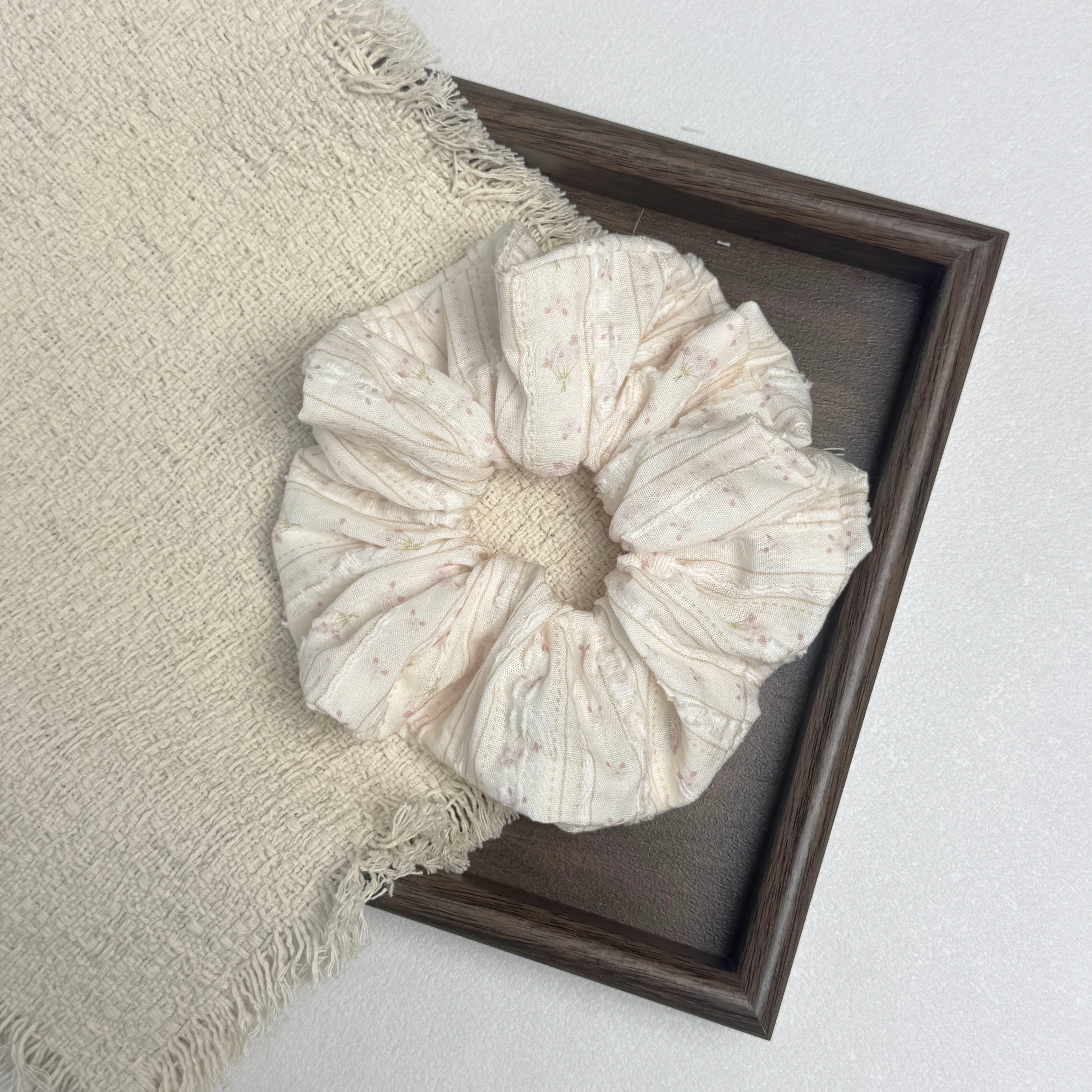 Cream floral bouquet print scrunchie flat-laid across a dark brown wooden tray and a frayed cream woven fabric with tasseled edges, white textured surface.
