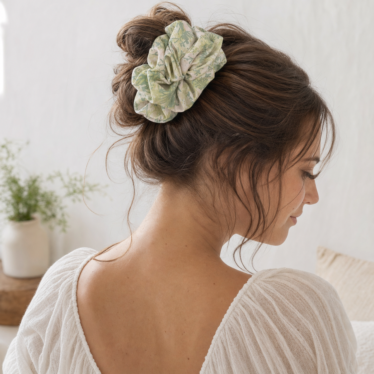 Woman with brown hair in a high messy bun secured by a cream green floral scrunchie, back and side profile, white crinkle-gauze V-back blouse. Small fern in white pot on wood stump in background.