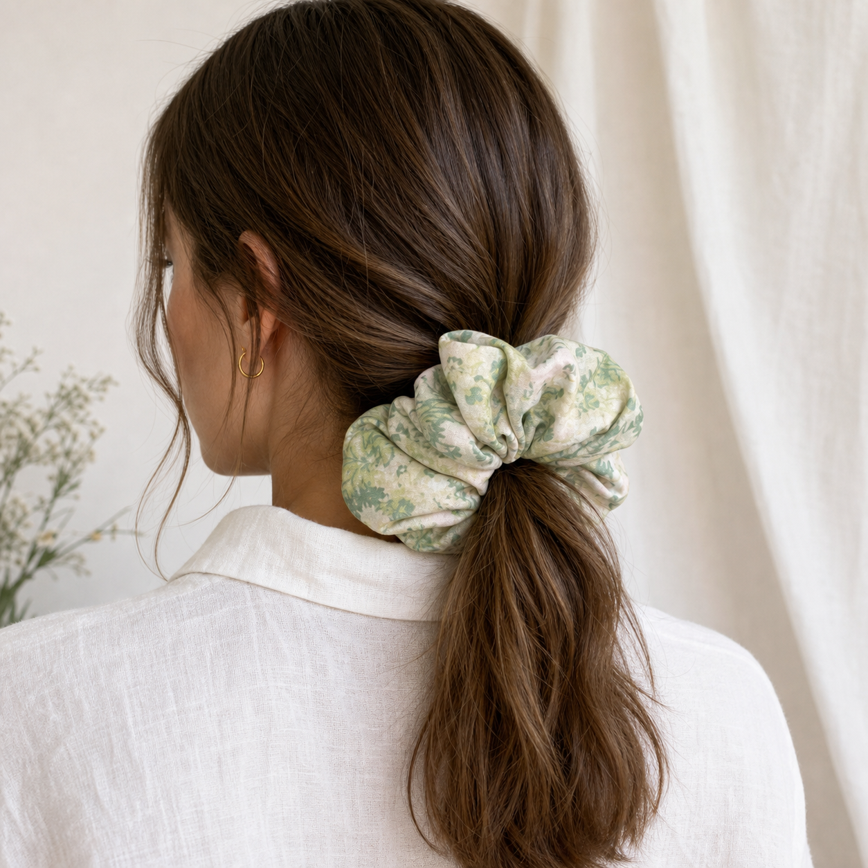 Woman with brown hair in a low ponytail secured by a cream green floral print scrunchie, rear view, white linen shirt, gold hoop earring. Soft white curtain and green foliage in blurred background.