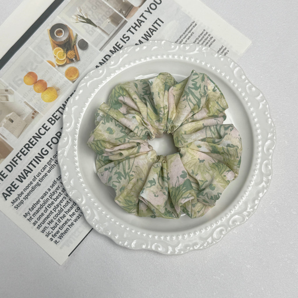 Cream green floral print scrunchie placed on a white ceramic plate with embossed scalloped rim, resting on a magazine page featuring fruit and spice photography, white grainy surface.
