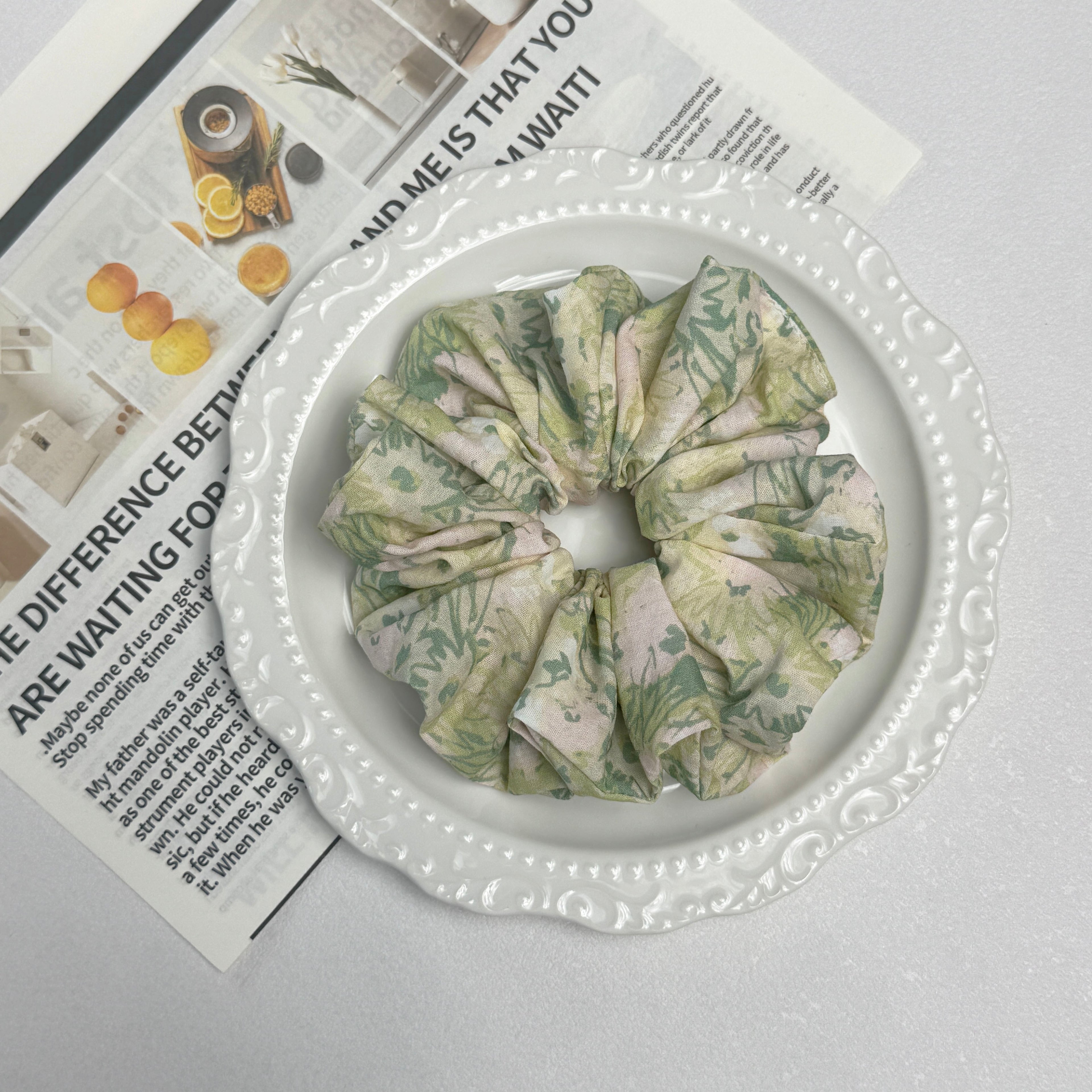 Cream green floral print scrunchie placed on a white ceramic plate with embossed scalloped rim, resting on a magazine page featuring fruit and spice photography, white grainy surface.