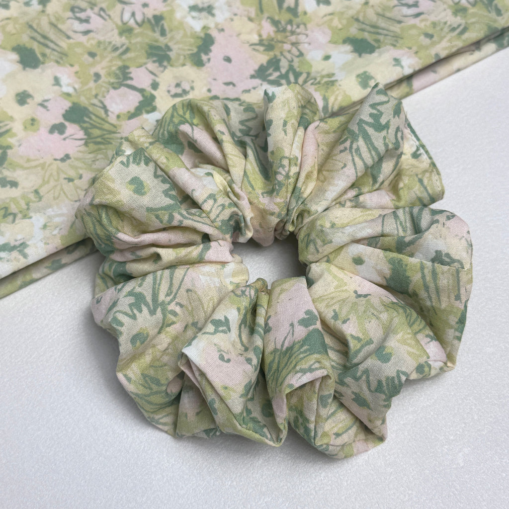 Cream green floral print scrunchie alongside a flat-laid matching fabric swatch showing the full watercolor botanical pattern with sage, moss, blush, and pale yellow tones, white grainy surface.