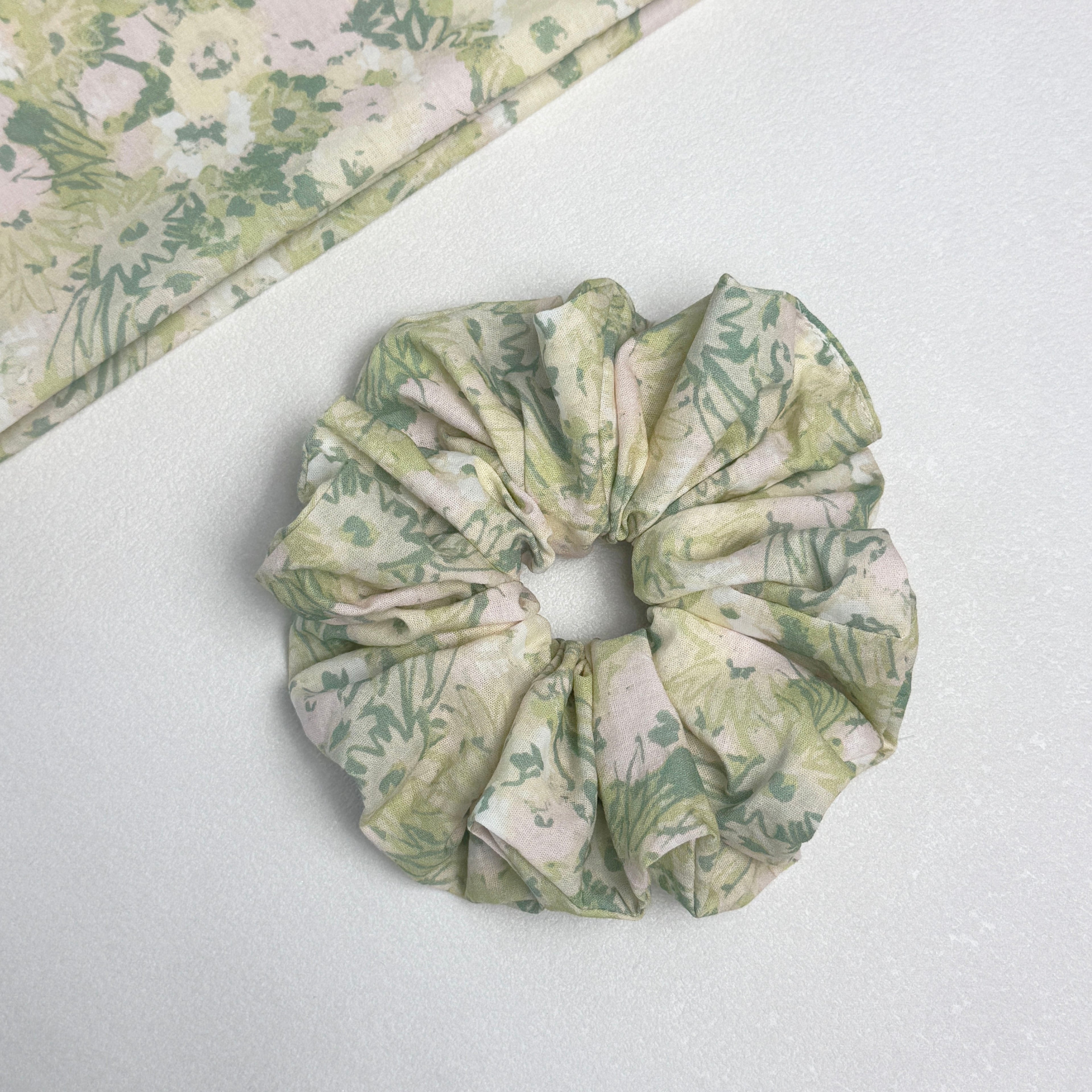 Cream green floral print scrunchie displayed alongside a matching folded fabric swatch showing the botanical print in sage, olive, and dusty pink tones, light grey pebbled surface.