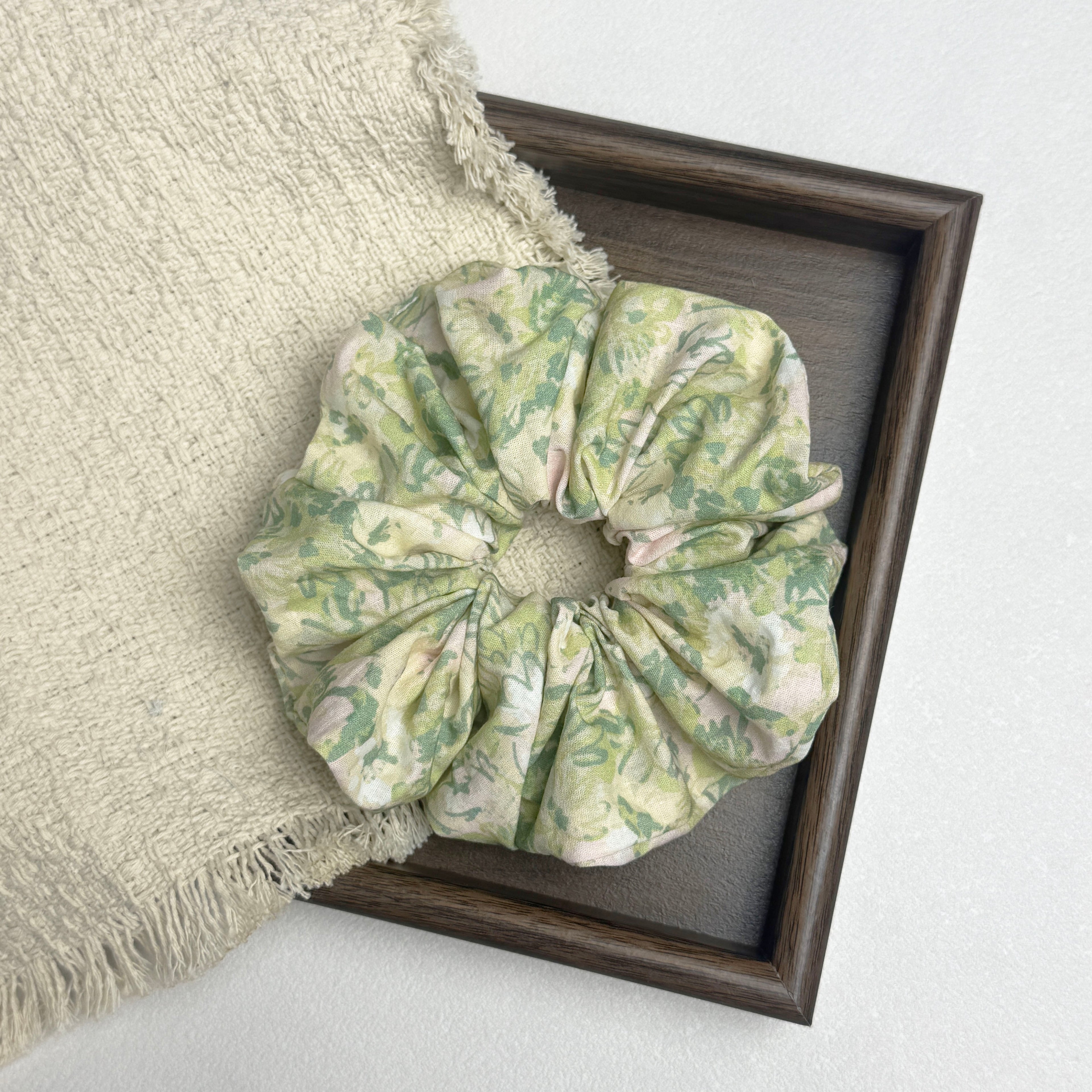 Cream green floral print scrunchie flat-laid across a dark brown wooden tray and a frayed cream woven fabric with tasseled edges, white textured surface.